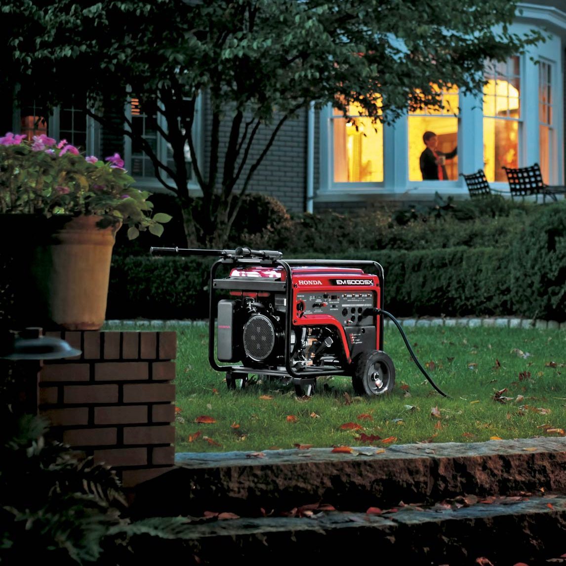 A portable generator is placed on a lawn in the foreground, with a large house in the background displaying illuminated windows. Inside the house, two people are visible through the windows, possibly engaged in conversation. The scene is set during the evening.