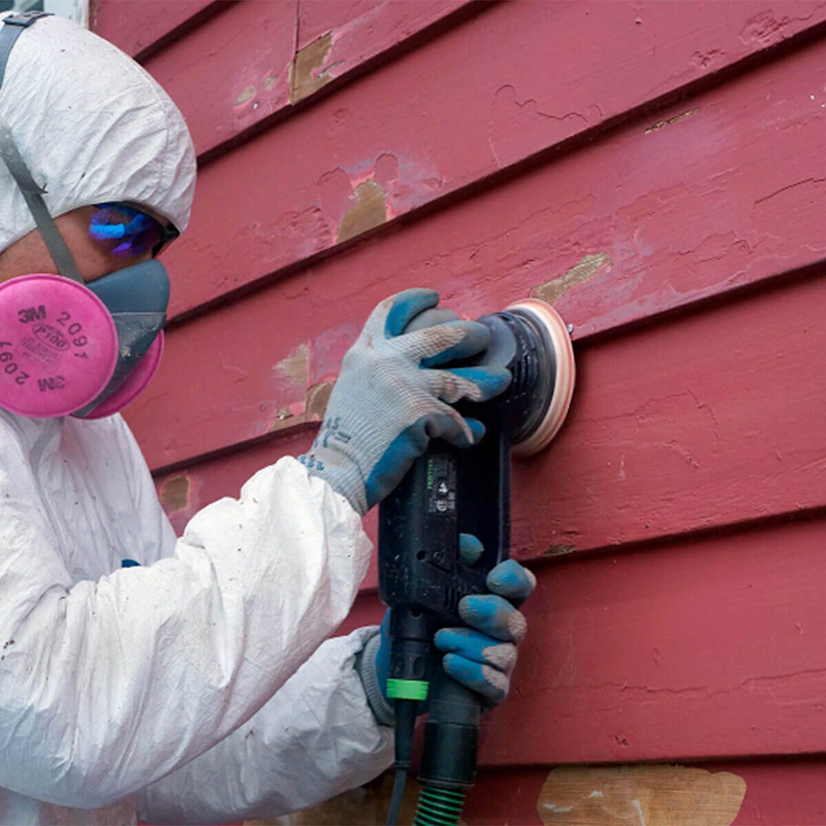 man sanding peeling paint on red house