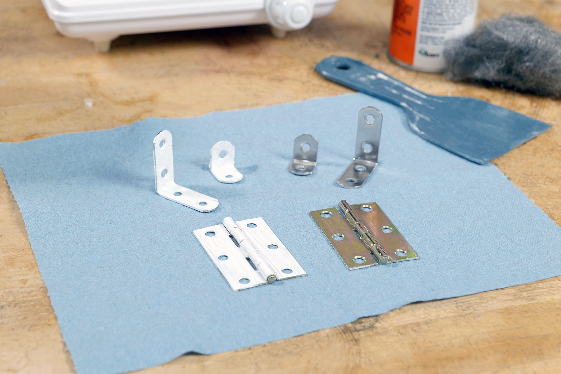 Different types of metal brackets and hinges are arranged on a blue cloth. A putty knife, a container, and other tools are visible in the background on a wooden surface.