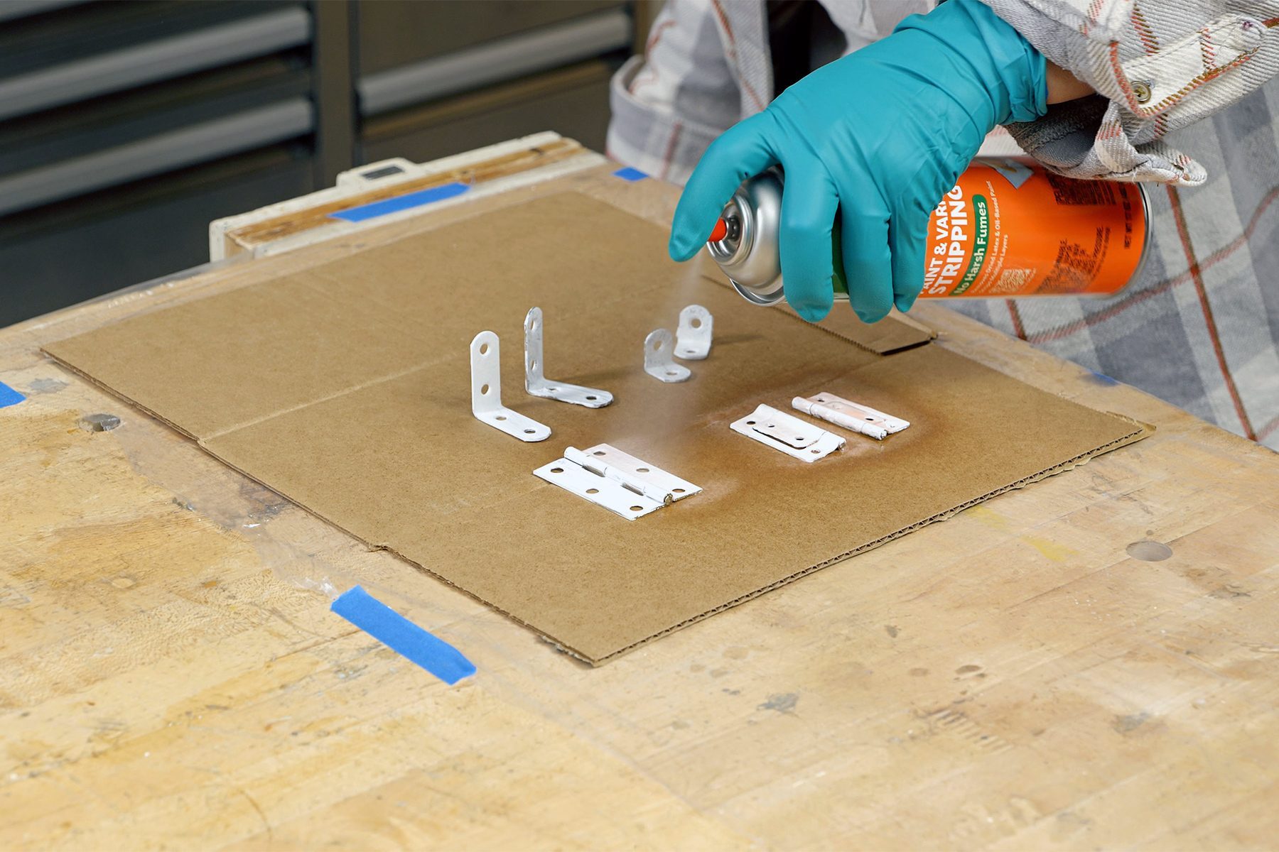 A person wearing teal gloves sprays paint from an orange can onto several metal brackets and small metal plates positioned on a piece of cardboard on a workbench. Tools and blue tape are visible on the wooden surface surrounding the cardboard.