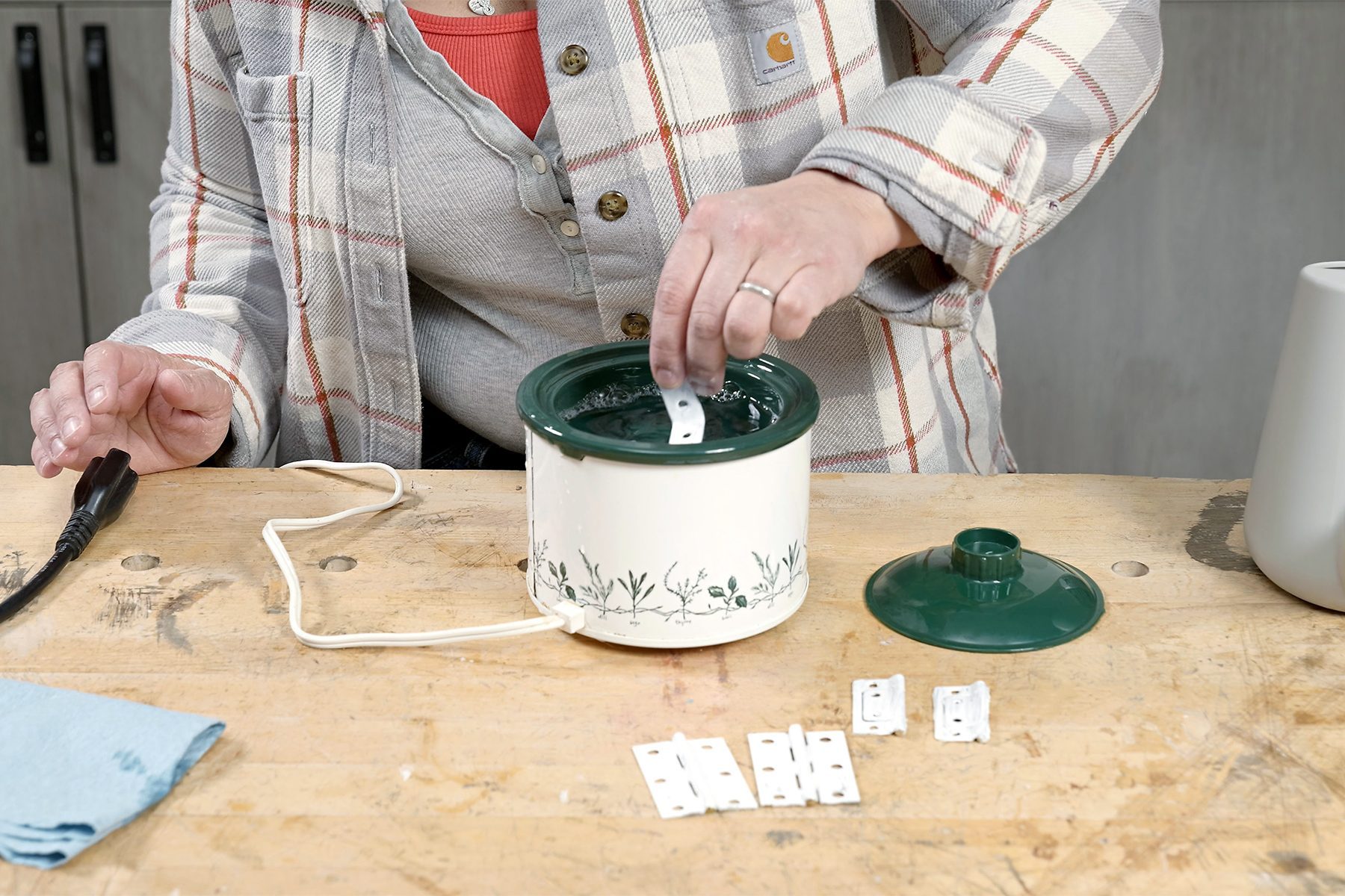 Person in a plaid shirt testing white strips in the green and white pot of a wax warmer on a wooden table, with spare strips and a lid beside the warmer.