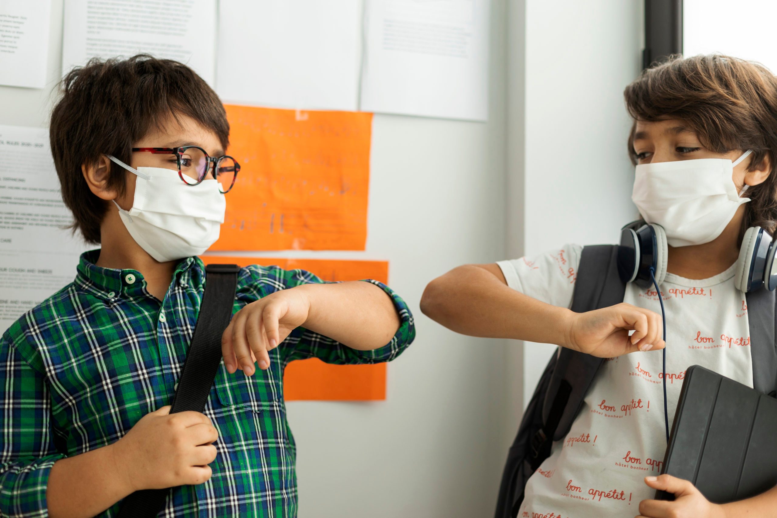 Boys wearing masks giving elbow bump while standing against wall in school