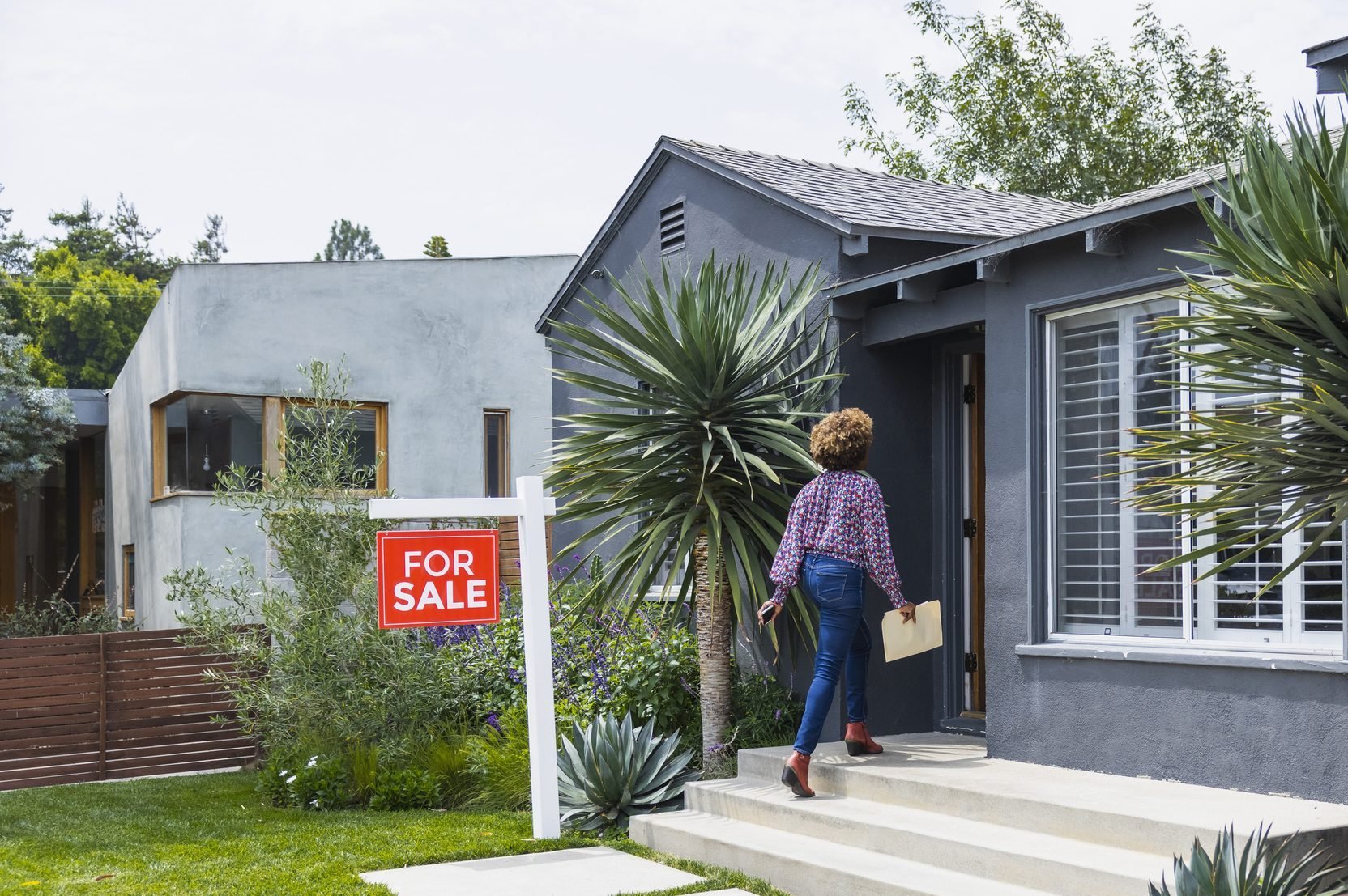 Female real estate agent carrying document while walking into house with a for sale sign out front