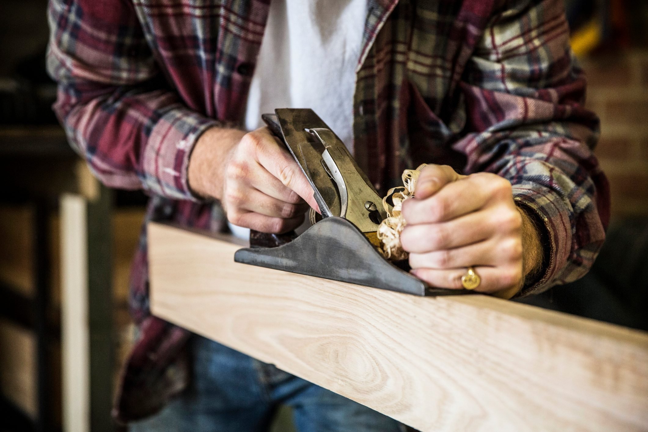 Close up of man working standing in a woodworking workshop, using hand plane on plank of wood.