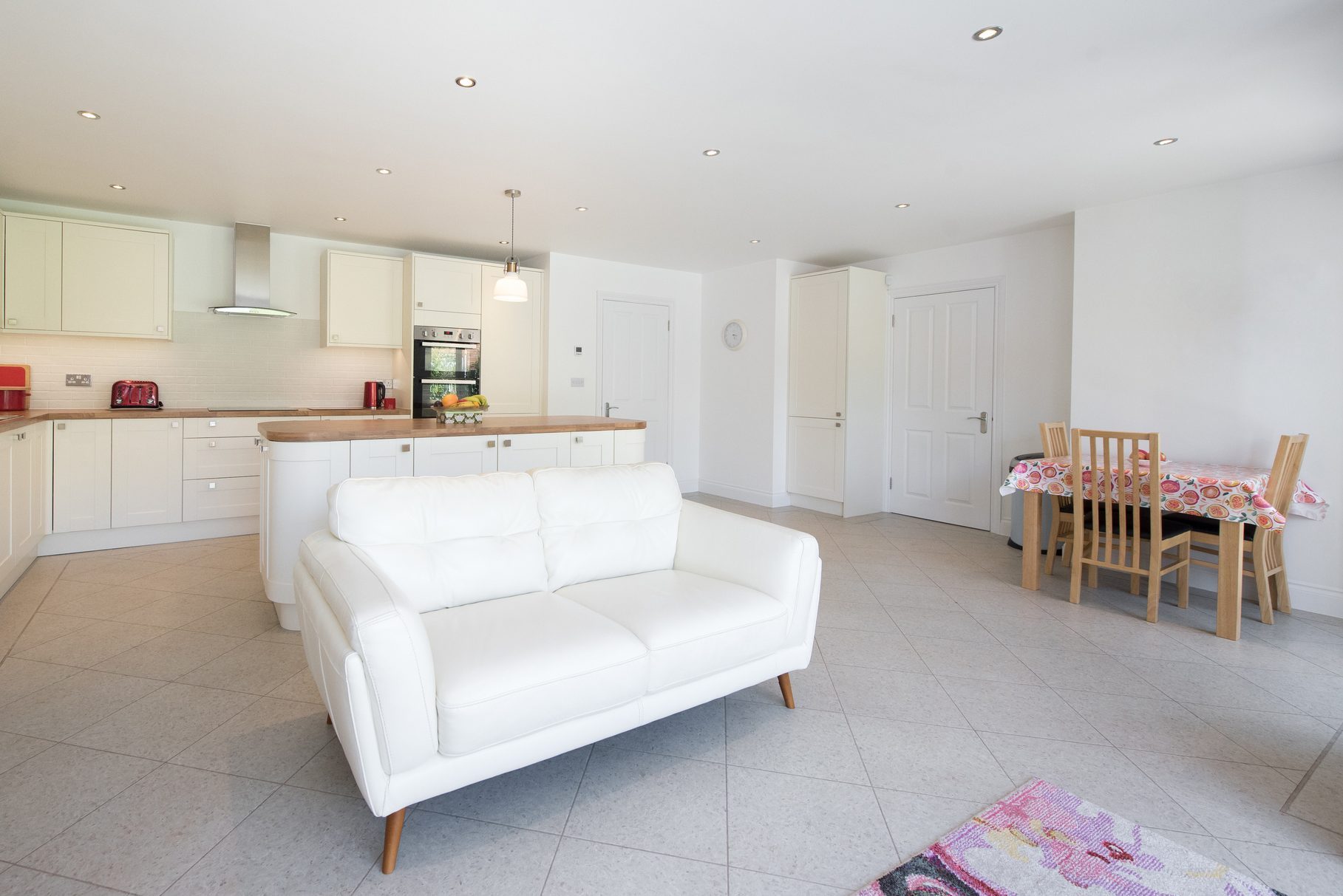 A general interior view of cream coloured a fitted kitchen diner with rounded corner cabinets, centre island, wooden worktop, leather sofa, round spot light, down lights and dining table and chairs