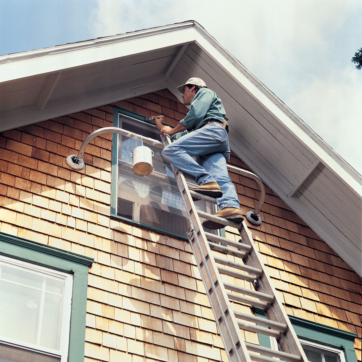A person in a green shirt and jeans stands on a tall ladder, painting the upper exterior of a house with wood siding. They are using a paintbrush and a bucket of paint attached to the ladder. The house features a gable roof and green-trimmed windows.