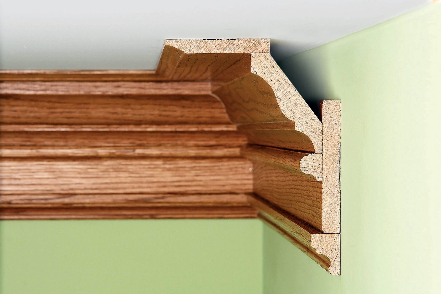 Close-up of a finely crafted wooden crown molding installed at a ceiling corner, with detailed profiles and layered sections. The molding is a light brown wood tone contrasting against the light green wall and white ceiling.