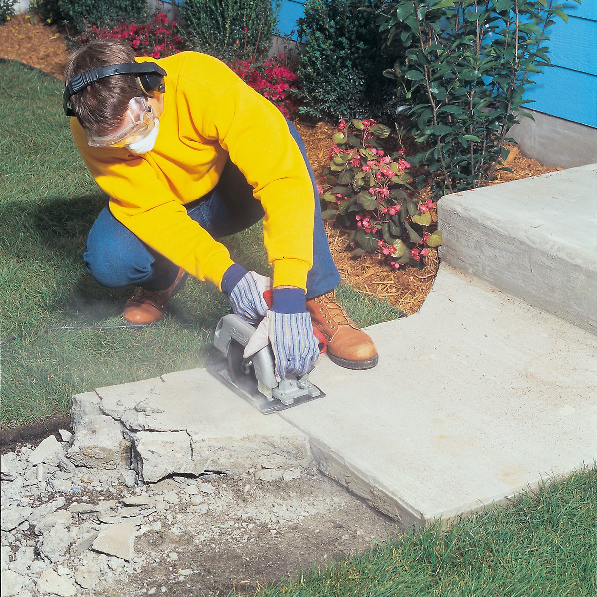 A person wearing protective glasses, ear protection, gloves, and a yellow jacket is using a circular saw to cut through a concrete slab next to a garden bed with green plants and flowers. The person is kneeling on one knee for better control of the saw.