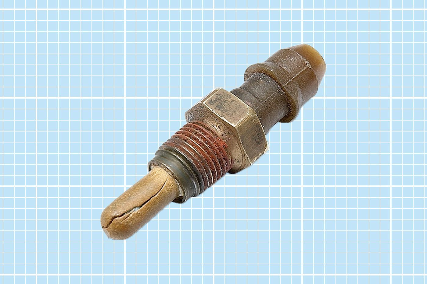 A worn, crack-filled engine ignition glow plug against a light blue graph paper background. The metallic component shows signs of heavy use, including rust and discoloration, and is used in diesel engines to assist in starting under low temperatures.