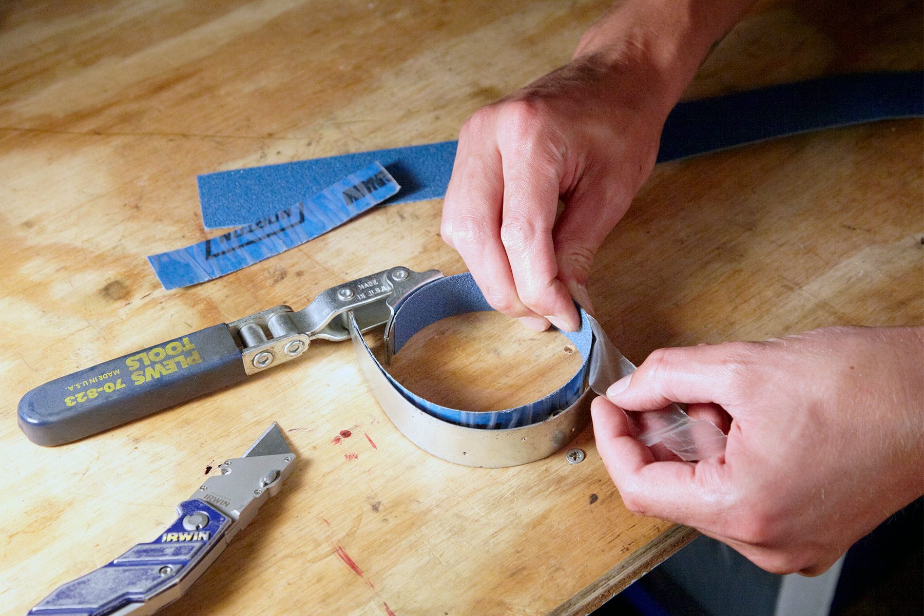 A close-up image of hands working on a wooden surface with tools. One hand is gripping a piece of blue sandpaper, while the other peels back a layer from it. A utility knife and tape measure are on the table nearby.