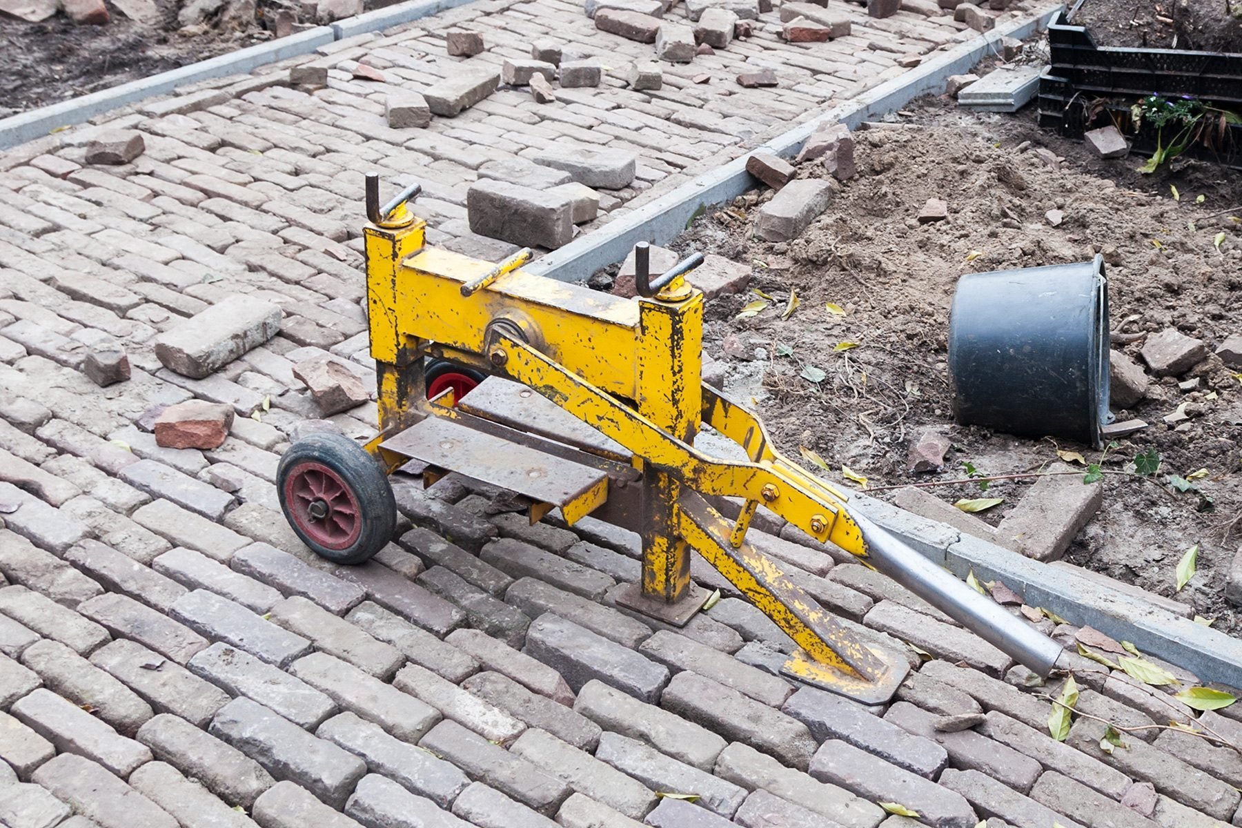 A yellow brick cutter on wheels is placed on a partially paved brick road under construction. The surrounding area shows scattered bricks and earth, with metal rails laid out to guide the installation of the bricks.