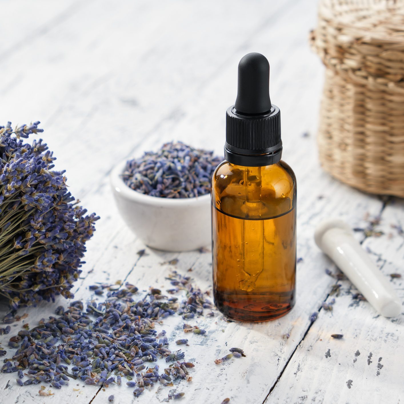 Dropper bottle of lavender essential oil, sachet bags and bouquet of dried lavender flowers on white wooden table.