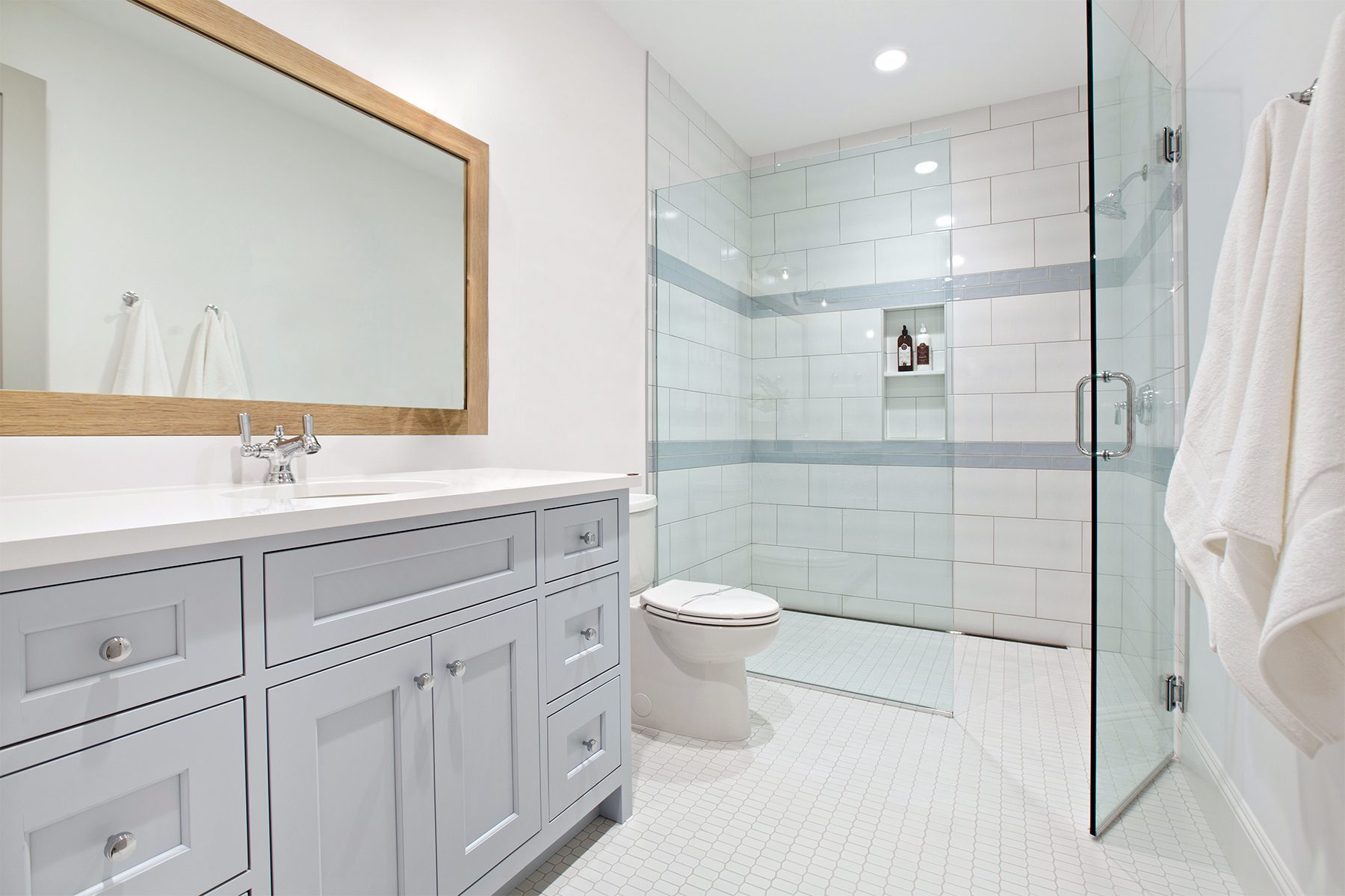 A modern bathroom featuring a white vanity with a wooden-framed mirror above it. There is a glass-enclosed walk-in shower with white and light gray tile. A white toilet is placed next to the shower. The floor is covered with small white tiles.