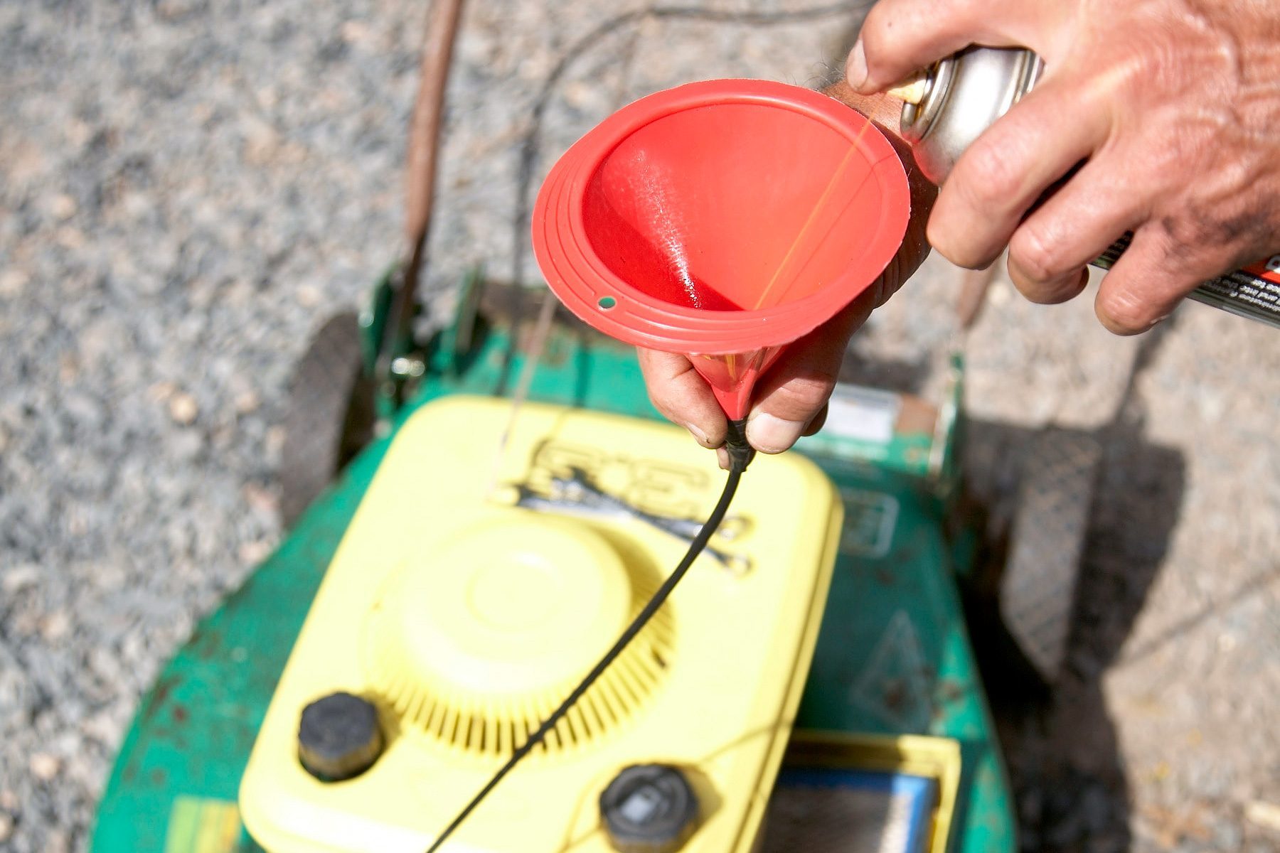A person is holding a red funnel and a can, likely adding oil or fuel to the yellow engine of a green lawnmower. The lawnmower is on a gravel surface.