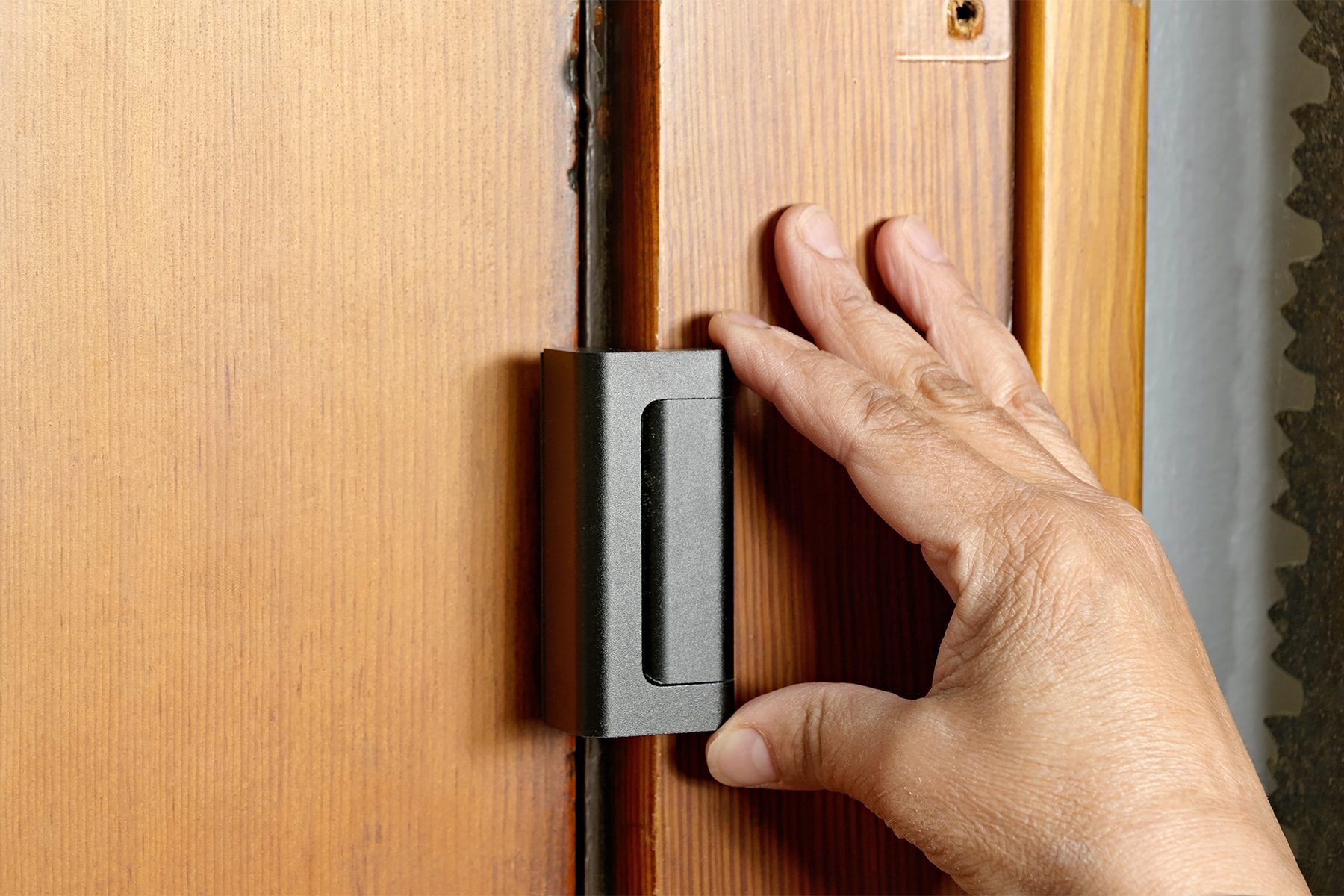 A hand is gripping a black sliding lock mechanism on a wooden door. The close-up shot captures only the hand and the door, focusing on the action of operating the lock.