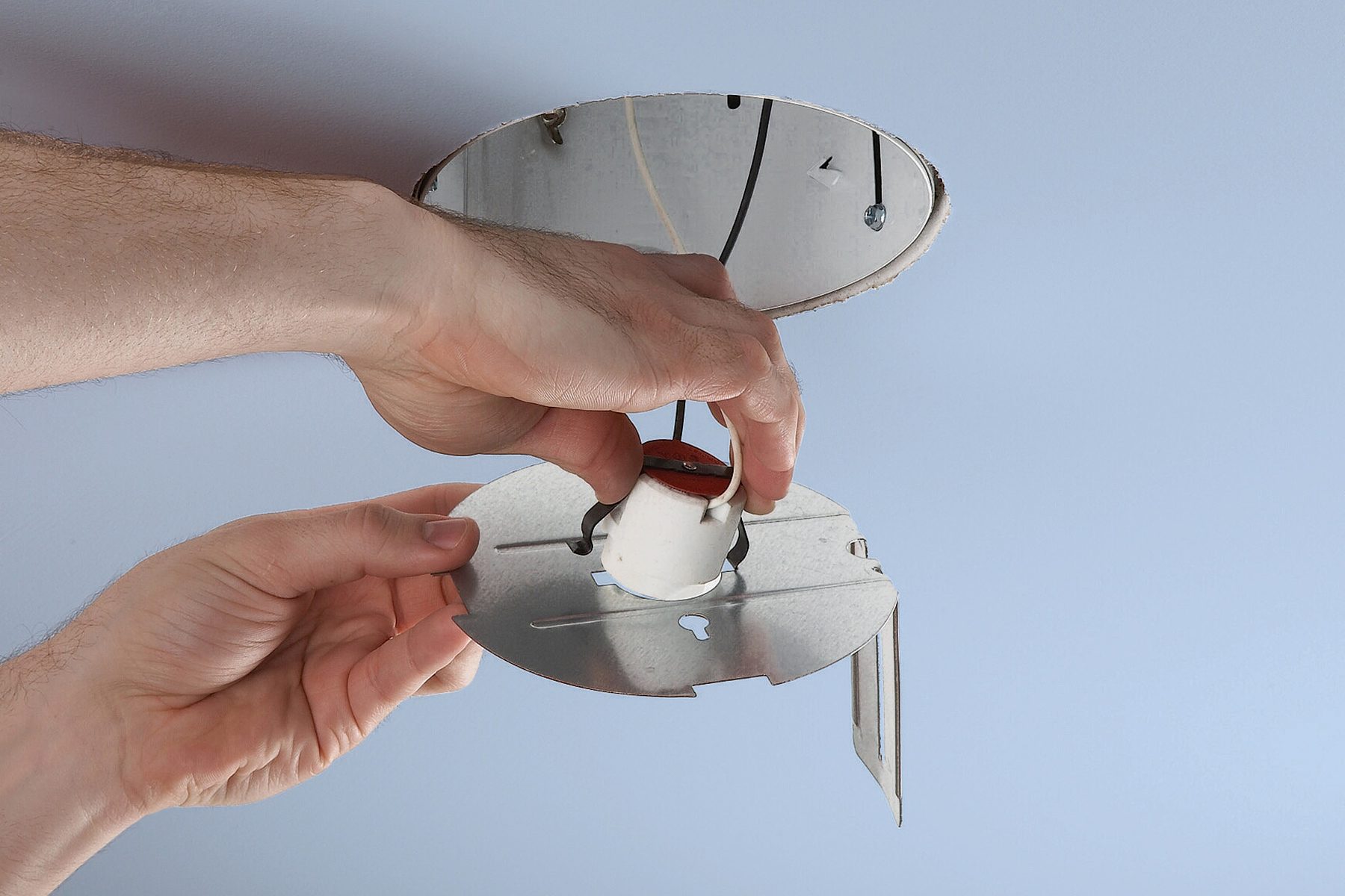 A person installing a light fixture in a ceiling. The person's hands are holding part of the fixture and aligning it into place. The fixture appears to be metallic with wires on the inside, and a bare ceiling opening is visible.