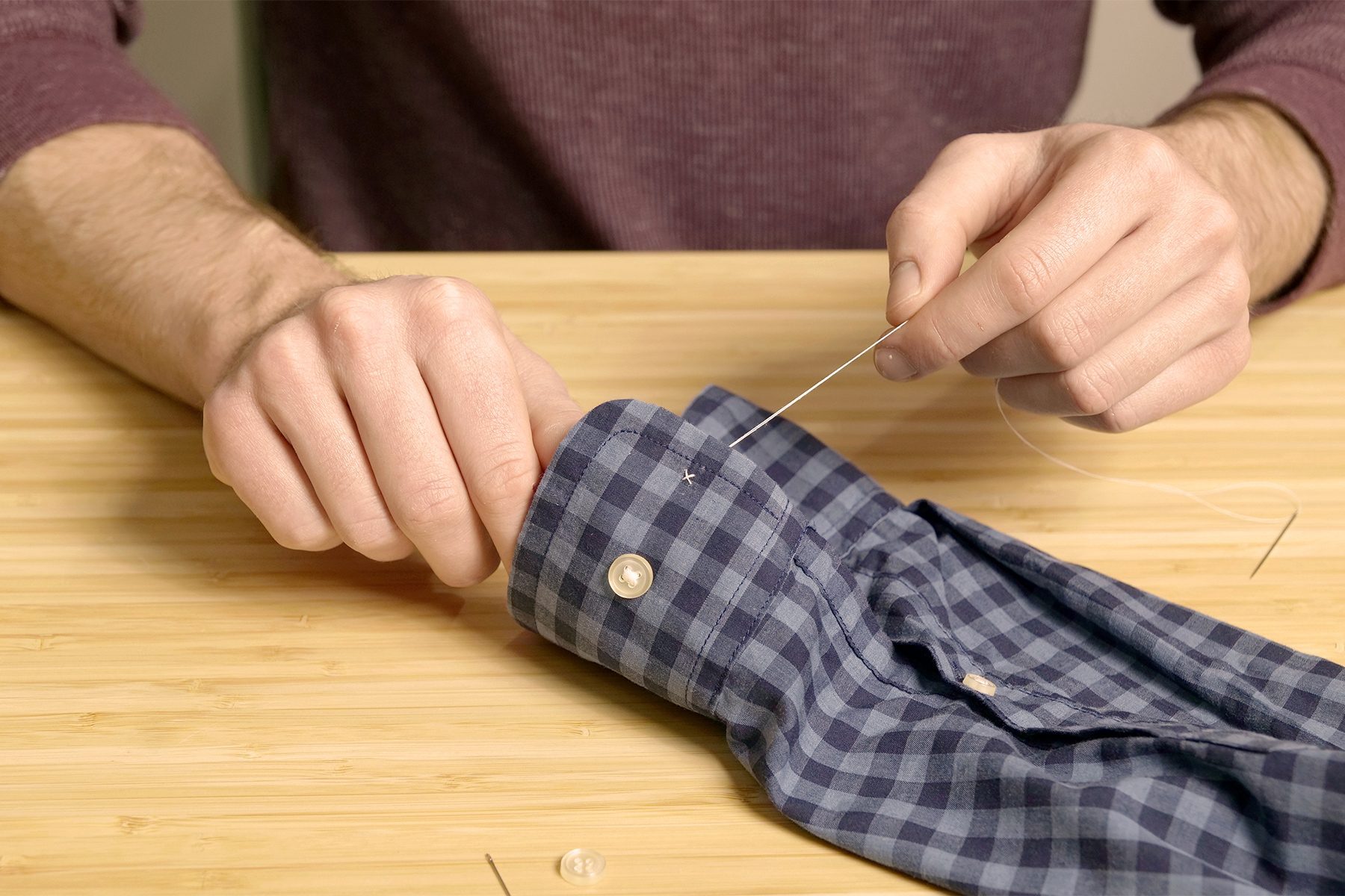 A person in a maroon long-sleeve shirt is sewing a button onto the cuff of a blue and white checkered shirt. The person is using a needle and thread, and the shirt is laid out on a light wooden surface.