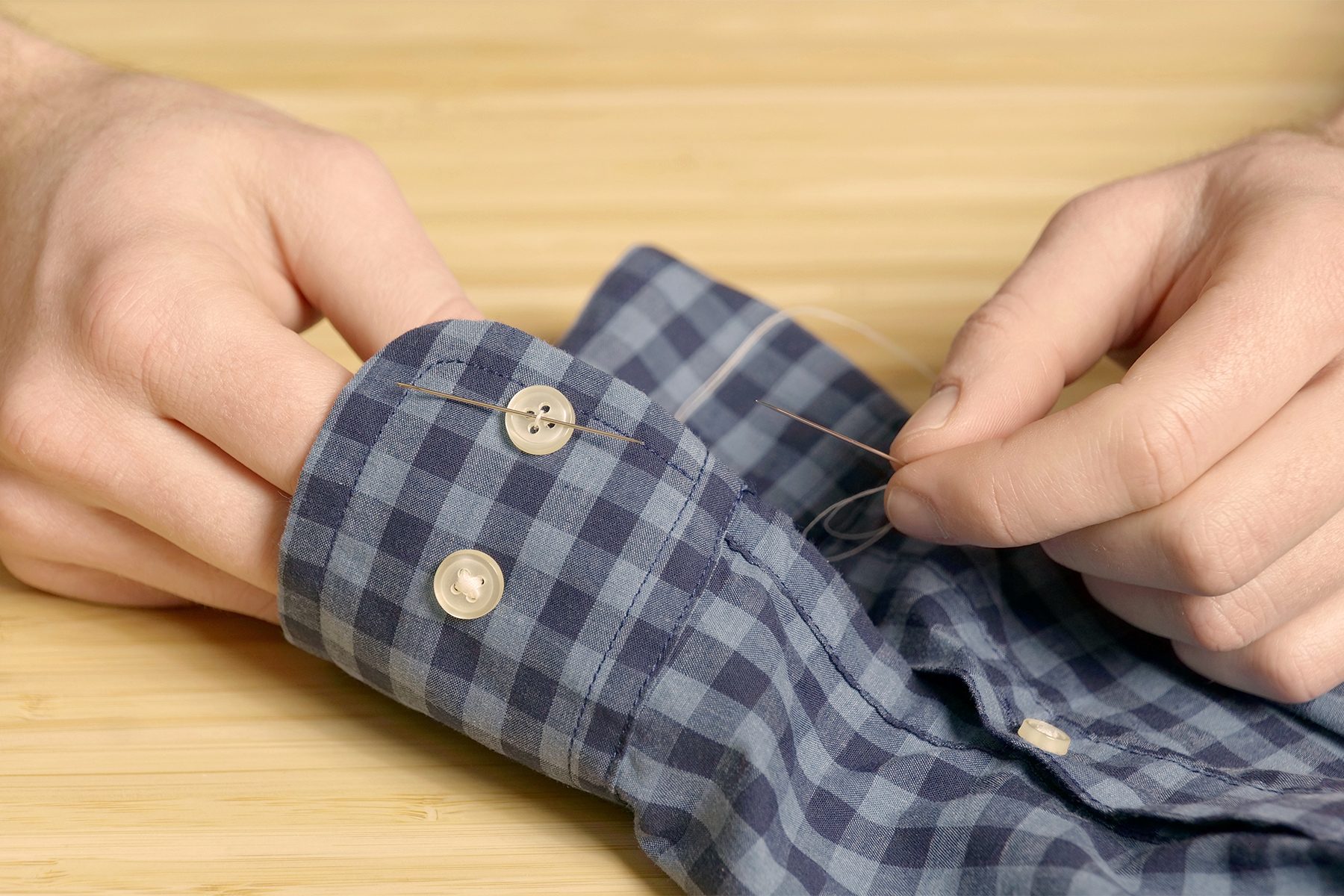 A person sewing a button onto the cuff of a blue and black checkered shirt. They are holding a needle with thread and working carefully on the wooden surface below. The cuff has another button already attached.