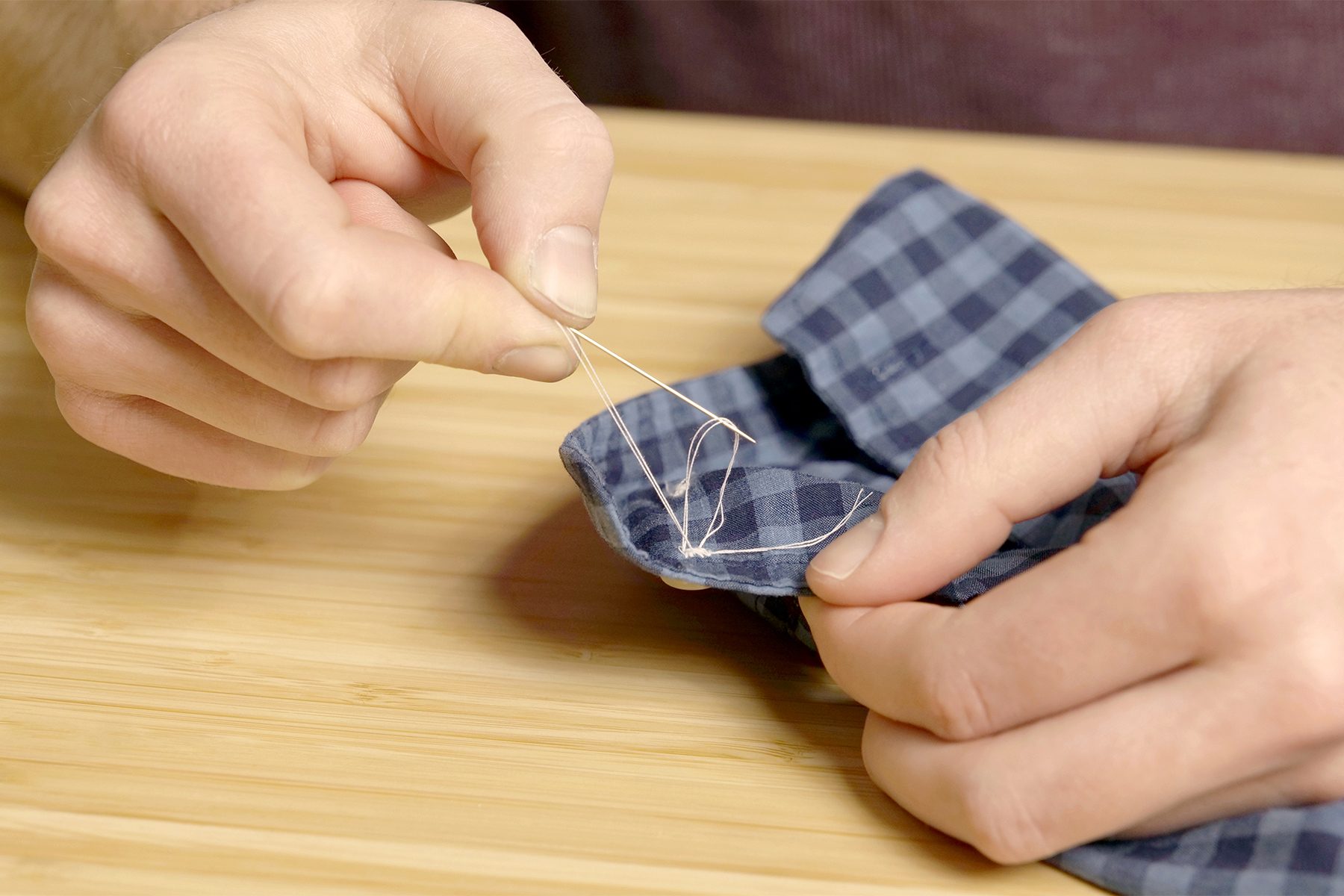 Close-up of hands sewing the blue and black checkered sleeve cuff of a shirt on a wooden surface. One hand holds the fabric while the other pulls a needle and white thread through the material.