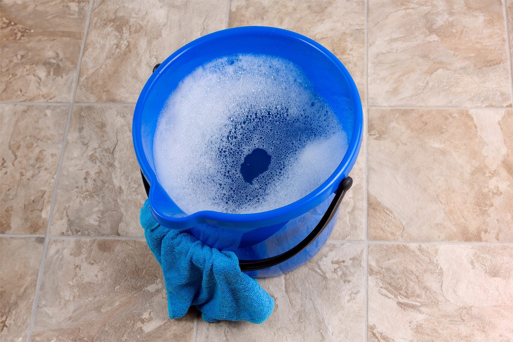 A blue plastic bucket filled with soapy water sits on a tiled floor. A blue cleaning cloth is draped over the edge of the bucket. The surrounding floor tiles have a beige and cream pattern.