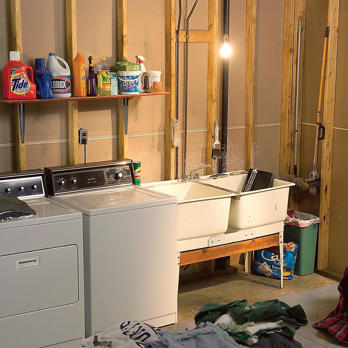 A laundry room with exposed studs and hanging light bulb. It has a washer, dryer, double sink, and shelves storing various cleaning products. Clothes are scattered on the floor. A garbage bin and laundry basket are in the background.