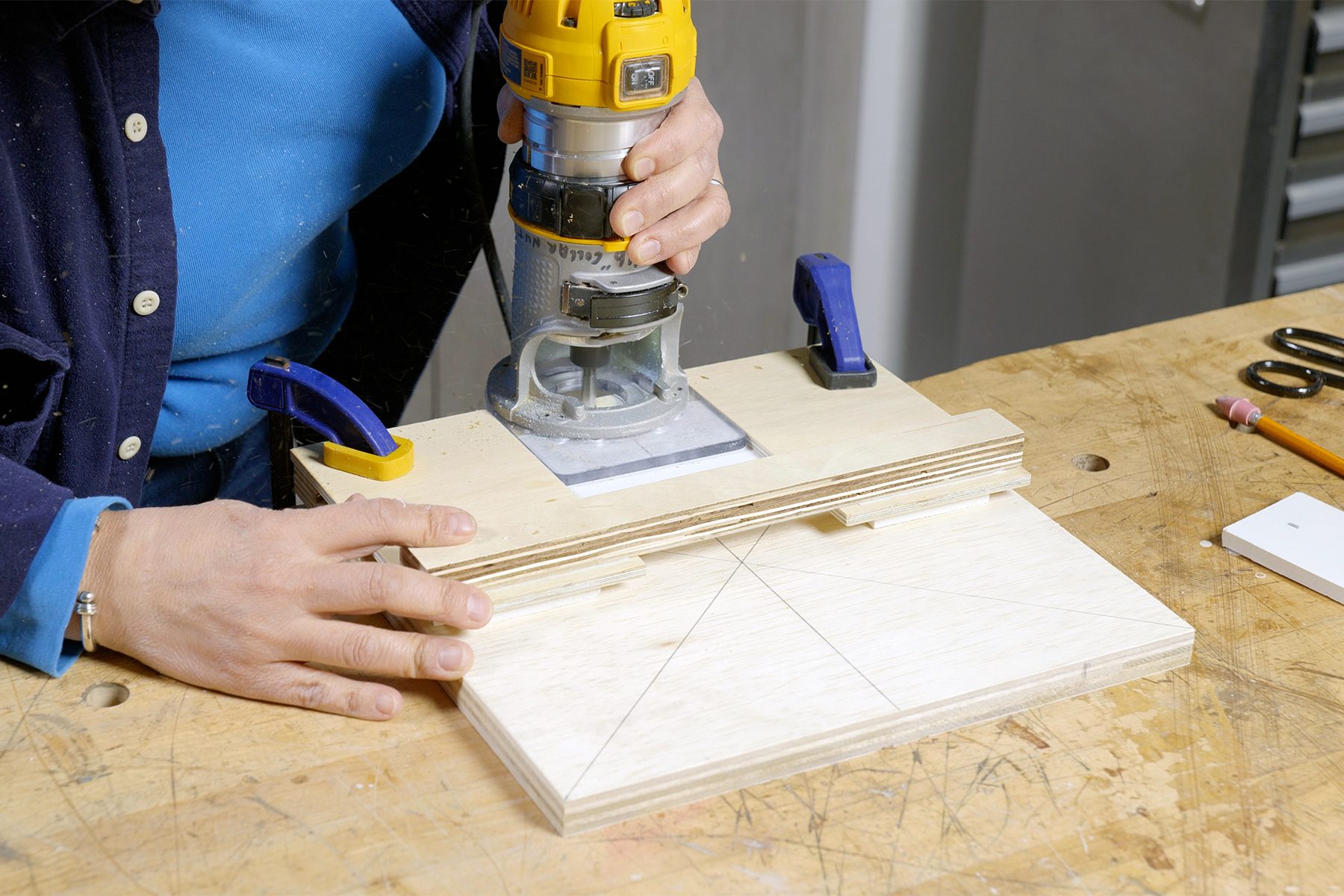 A person uses a yellow router tool on a piece of wood secured with clamps on a workbench. The wood has a penciled guideline of intersecting lines forming an "X." Tools are visible on the workbench in the background.