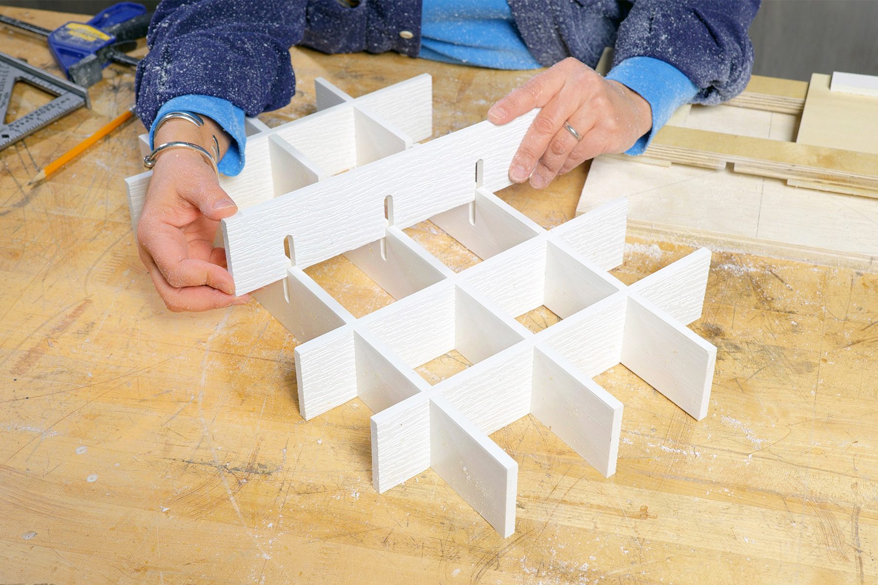 A person wearing blue sleeves assembling a white, grid-like organizer with multiple compartments on a wooden table. Various tools lie in the background, suggesting a crafting or woodworking environment.