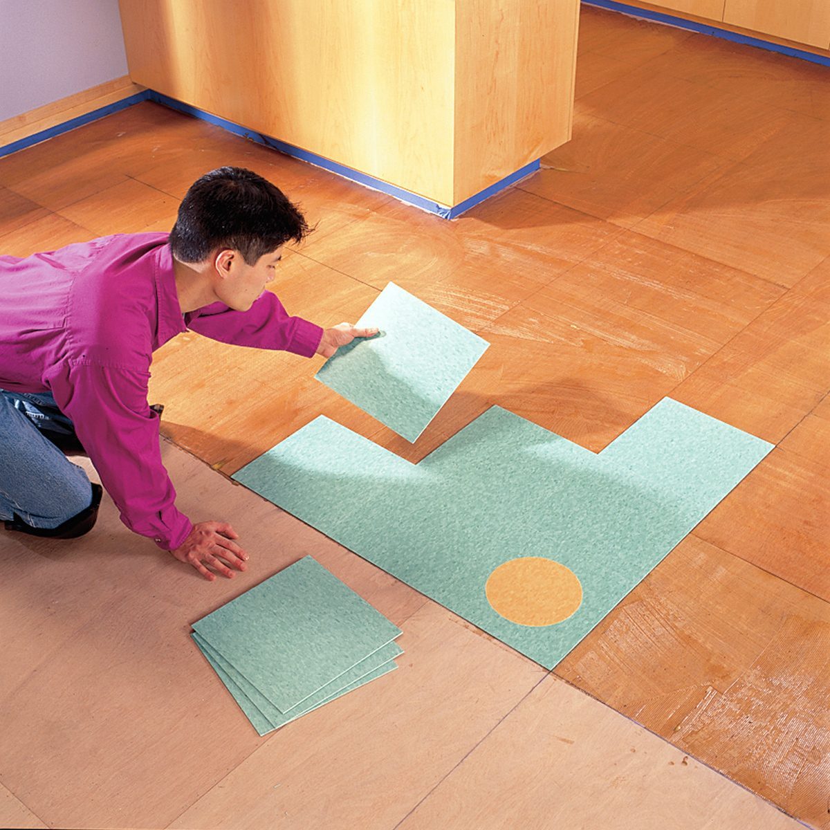 A person kneels on a wooden floor, fitting square tiles into place. The tiles are a light green color, with one tile featuring a circular tan design. Several more tiles are stacked nearby, ready to be installed. The setting appears to be an indoor space, possibly a home.