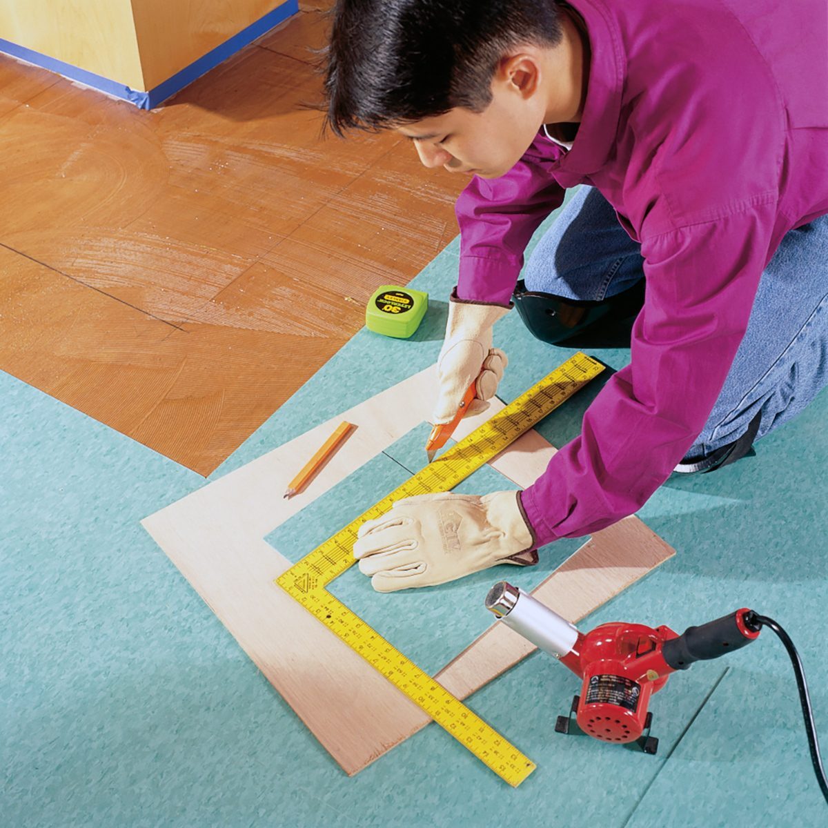 A person wearing gloves and a magenta shirt uses a measuring tape and straightedge to mark a piece of wood flooring. A green measuring tape, pencil, and red electric tool are on the floor nearby. The person is kneeling and working on a floor installation project.