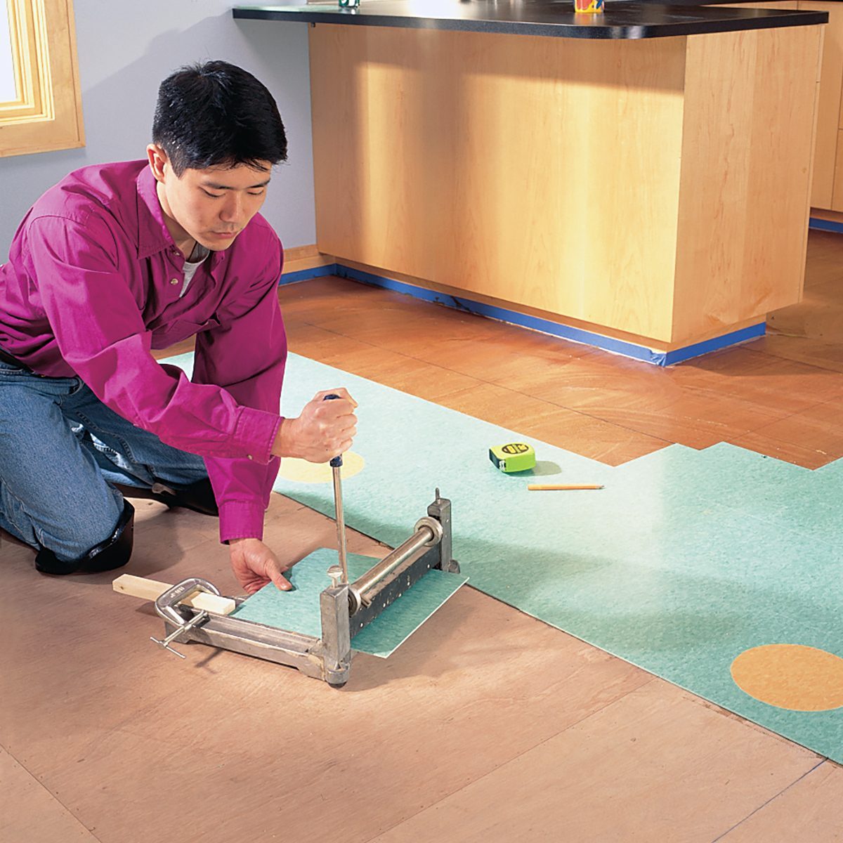 A person in a pink shirt and blue jeans kneels on the floor, using a tool to cut green vinyl flooring. A measuring tape, a piece of chalk, and a tool are nearby. The scene is set in a room with light-colored cabinetry and a wooden floor.