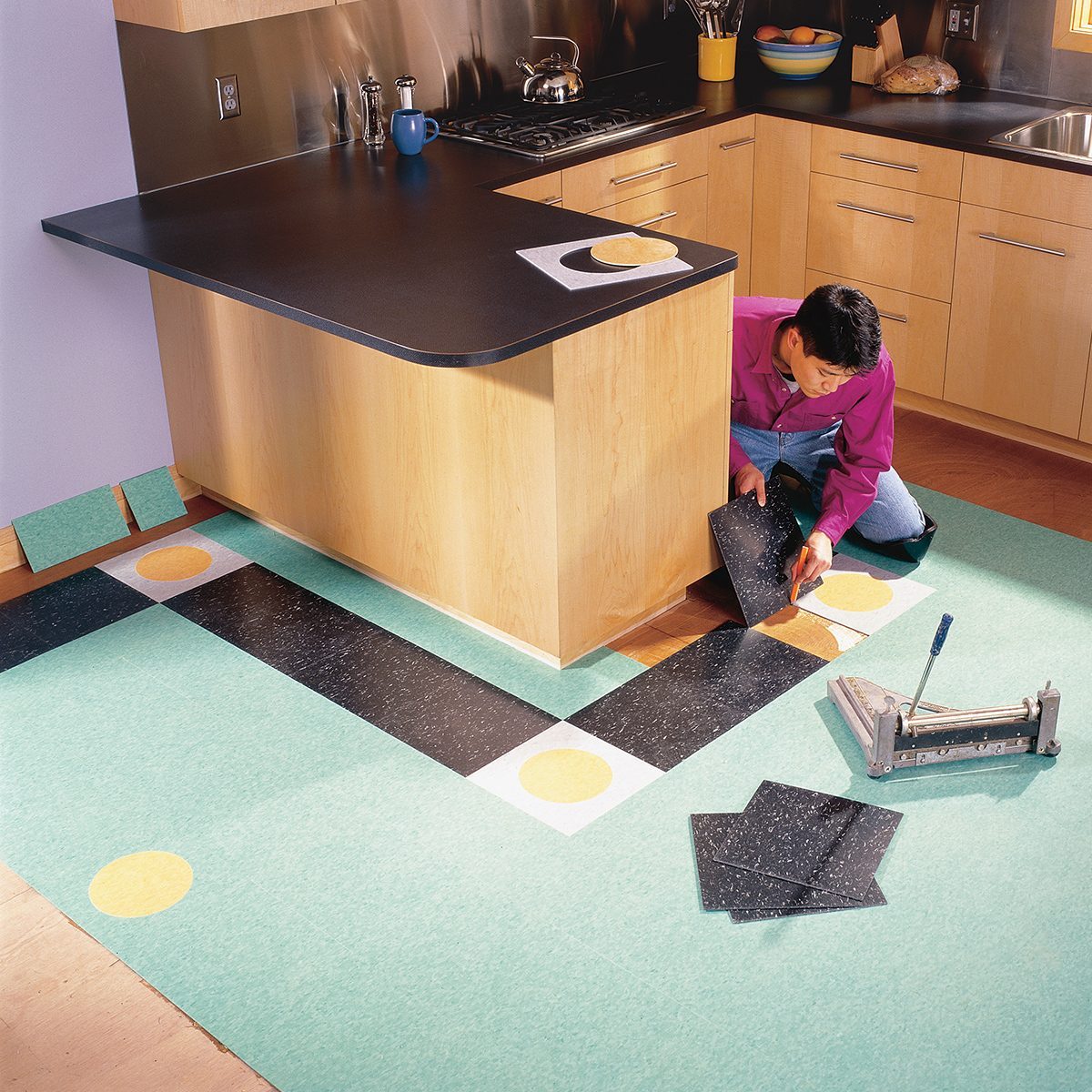 A person in a pink shirt is installing a new floor in a kitchen. The floor features a pattern of black and white sections with yellow circles. Tools and additional flooring materials are scattered around, and the countertops and cabinets are wooden.