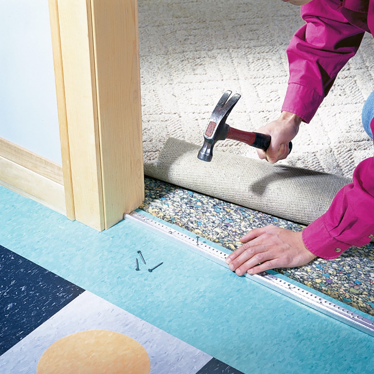 A person wearing a pink shirt is using a hammer to secure carpet to the floor with nails. The carpet is beige and they are working near a doorway. The floor nearby has a colorful pattern of blue, black, and yellow tiles.