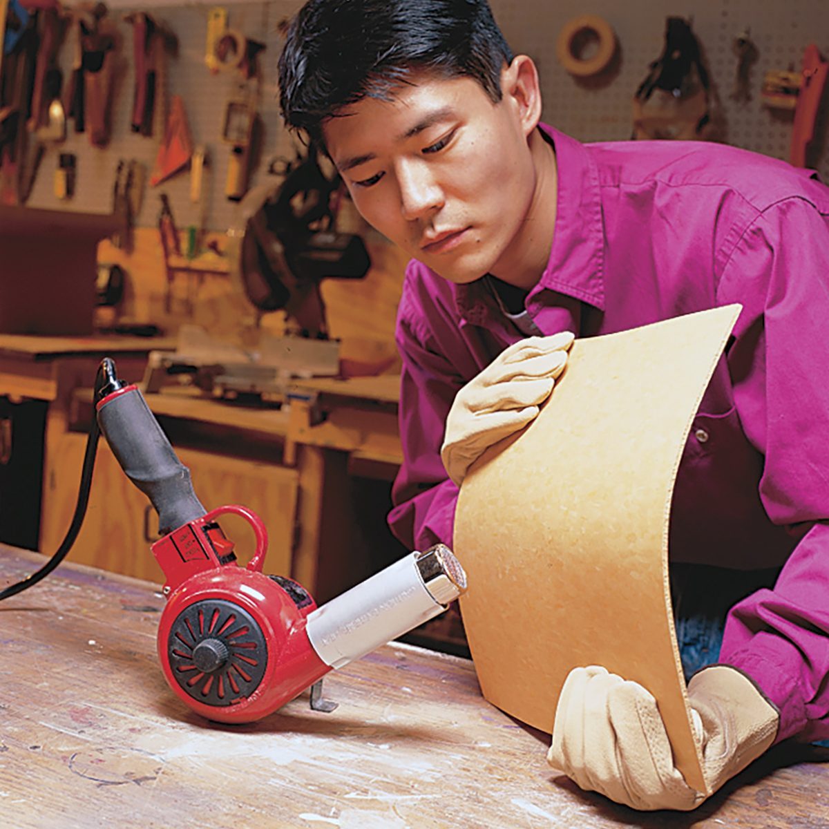 A person in a workshop wearing a magenta shirt and gloves uses a heat gun on a curved piece of material. The workshop background is filled with tools and equipment.