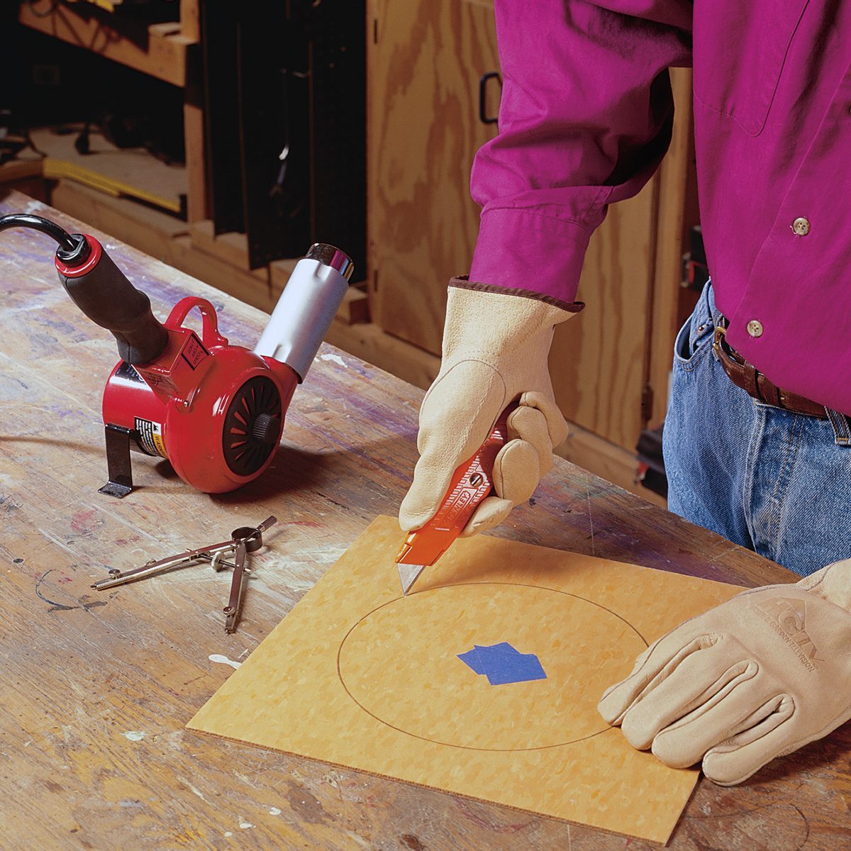 A person wearing gloves and a long-sleeved shirt uses a utility knife to cut along a circular outline on a piece of wood or paper. A red heat gun and other tools are on a wooden workbench nearby.