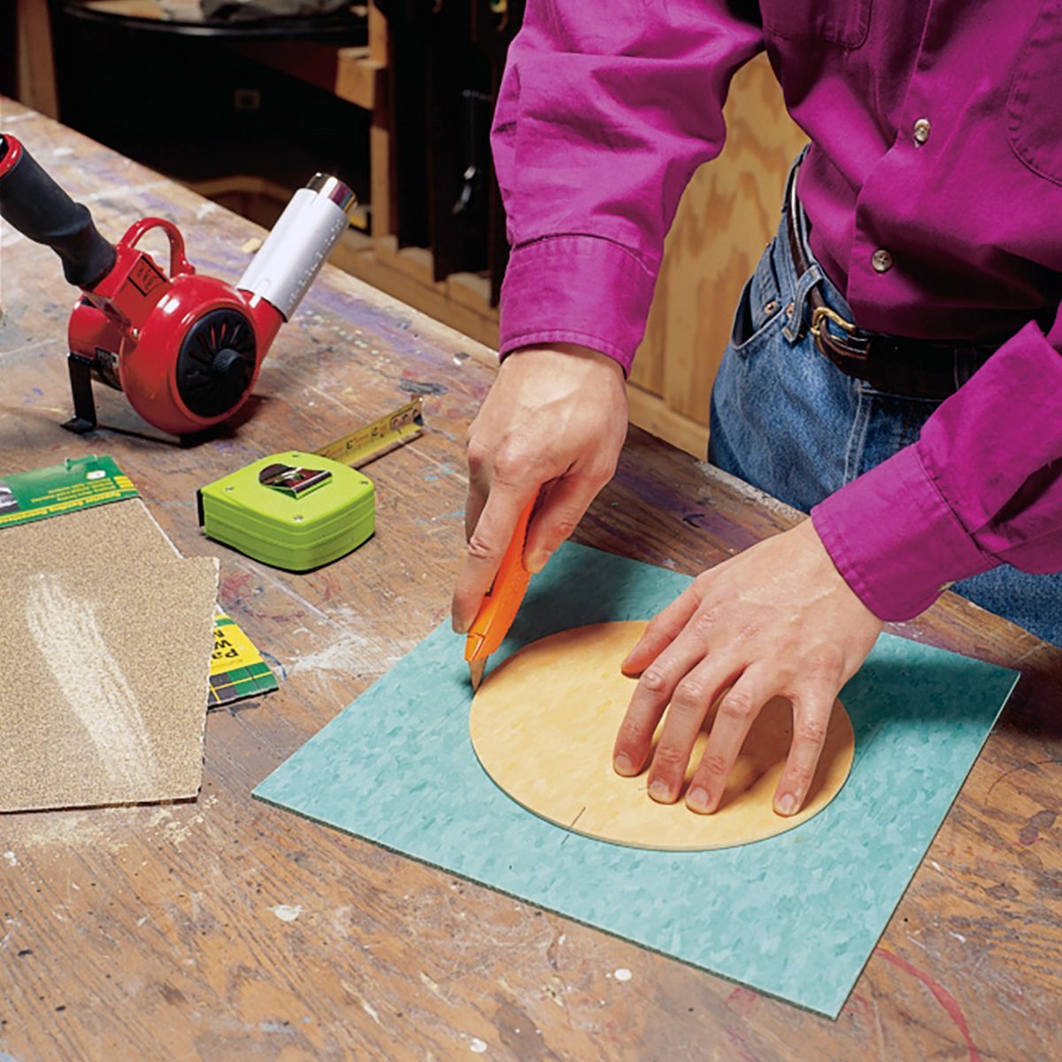 A person wearing a magenta shirt and blue jeans is using a utility knife to cut a circular shape out of a piece of green sandpaper on a wooden workbench. On the workbench, there’s a measuring tape, a package, and a red tool with a black handle.