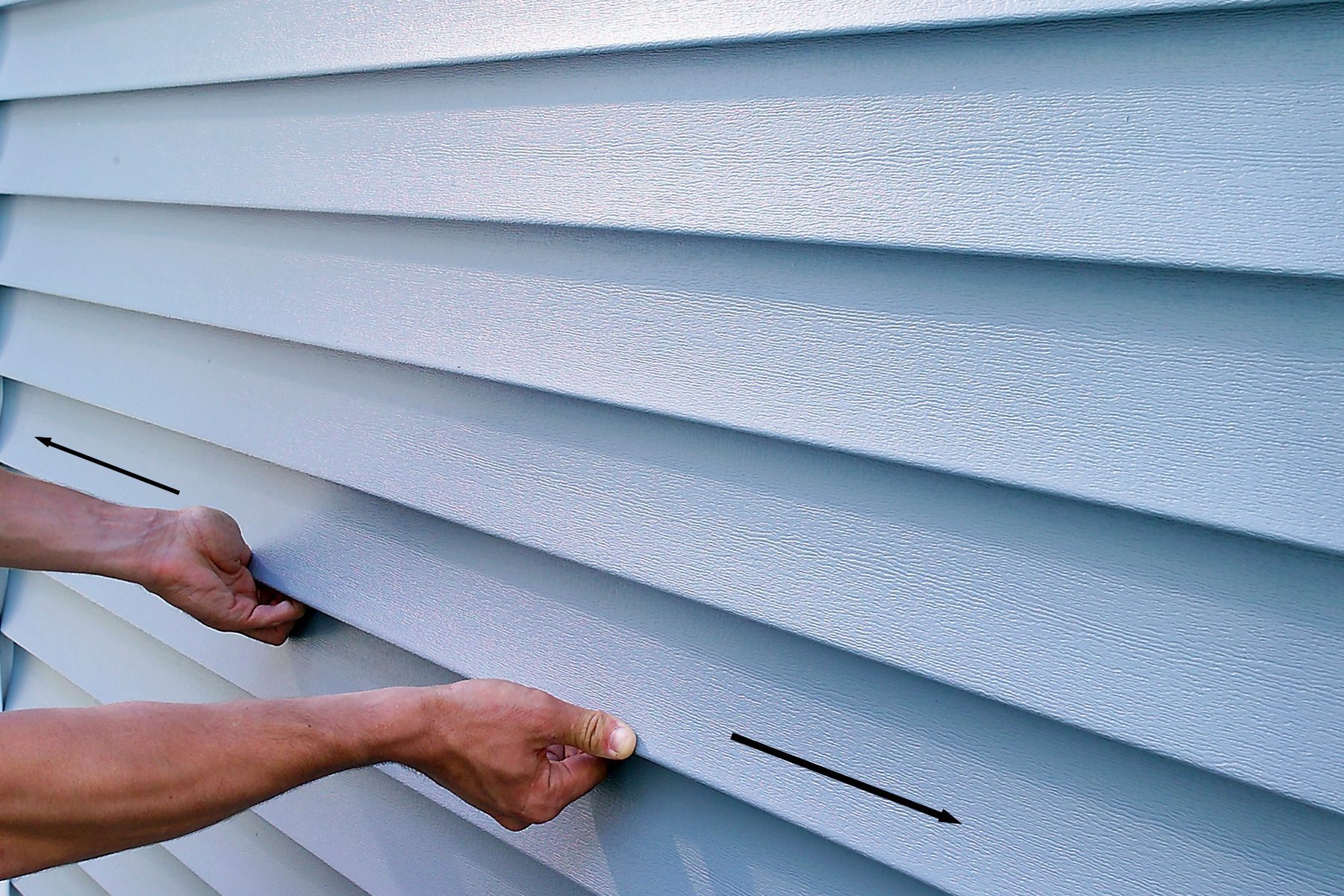 A person installs or adjusts horizontal vinyl siding panels on a building exterior. The image focuses on their hands placing a panel into position, showing the texture and alignment of several panels. The siding is light gray in color.