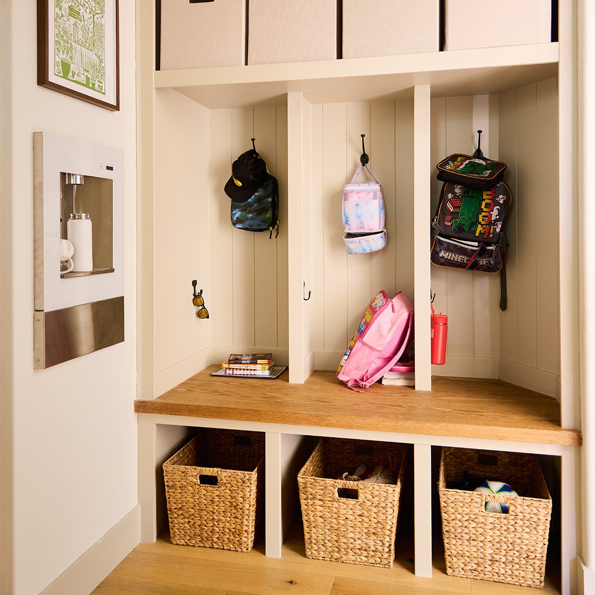 A neatly organized mudroom features three cubbies with hooks. Each cubby holds a backpack and some accessories. Below, there are three woven baskets for additional storage. The left wall has a mounted water bottle filler and a framed map above it.