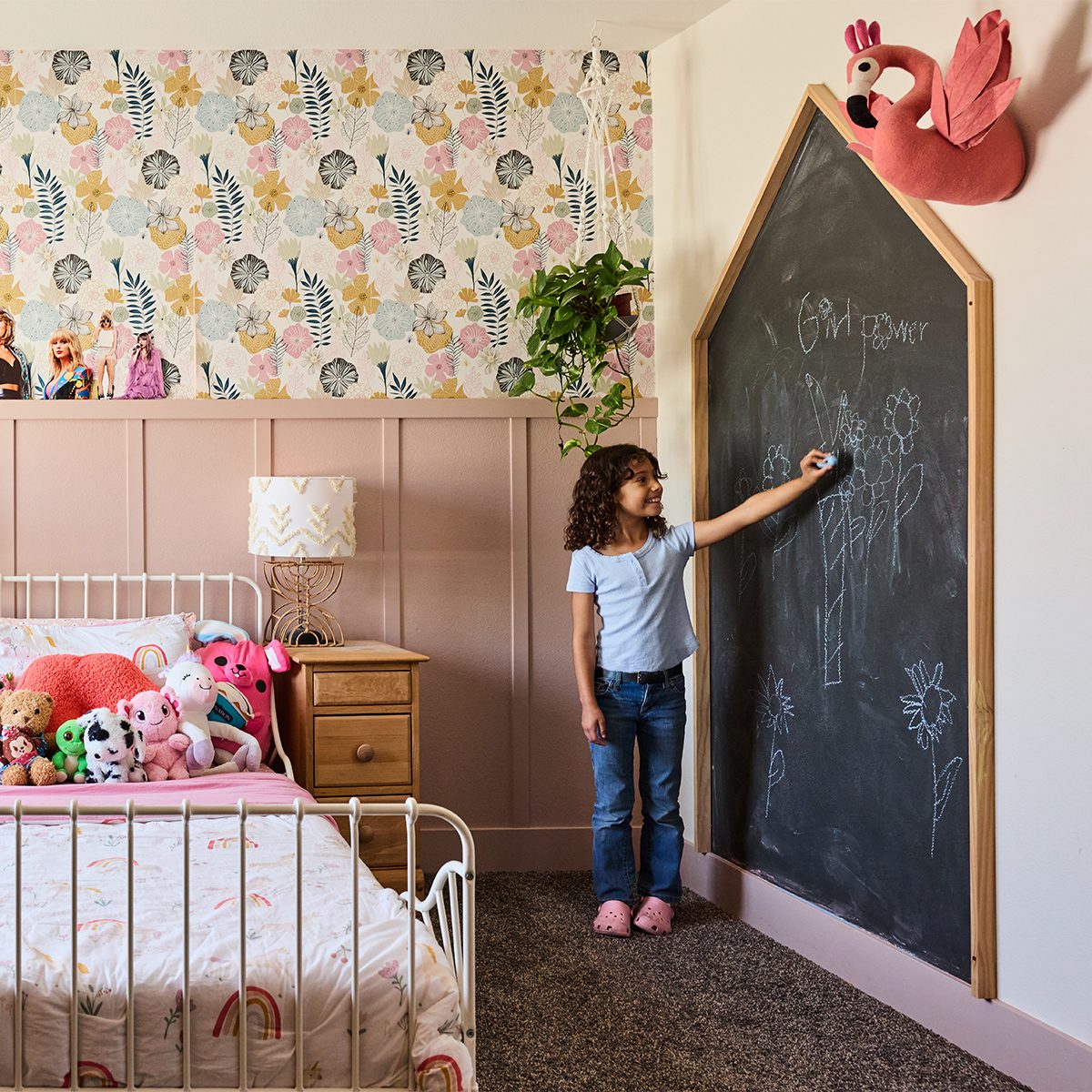 A young child stands at a large chalkboard shaped like a house, drawing with chalk in a child's bedroom. The room features floral wallpaper, a bed with stuffed animals, a nightstand with a lamp, and a flamingo wall decoration.