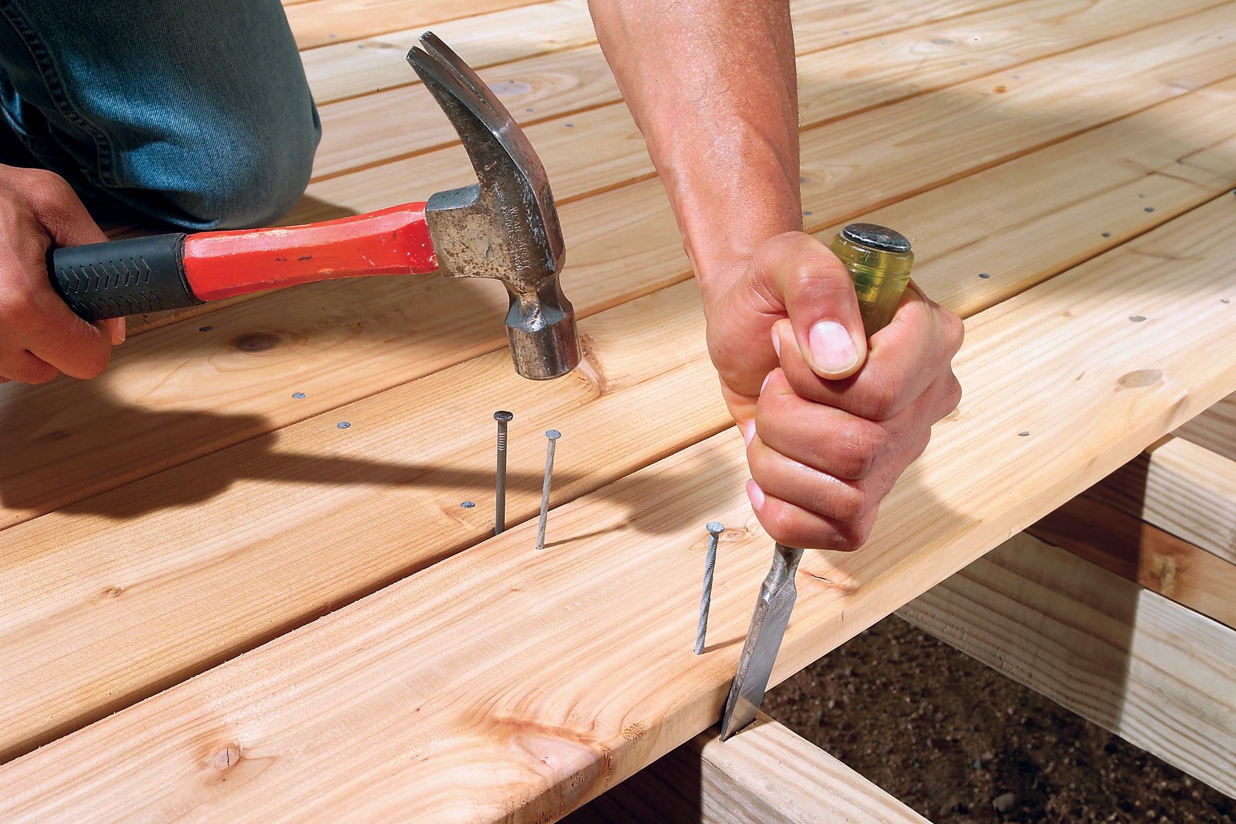 A person uses a hammer and a chisel to position and secure nails into a wooden deck. The person is wearing jeans and is working outdoors on a sunny day. The deck consists of neatly aligned wooden planks.