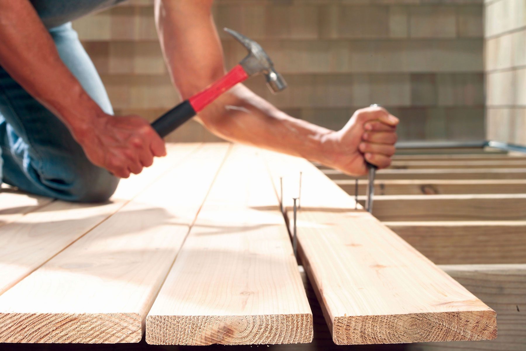 A person is using a hammer to drive nails into wooden planks. They are kneeling on the ground, and only their arms are visible.