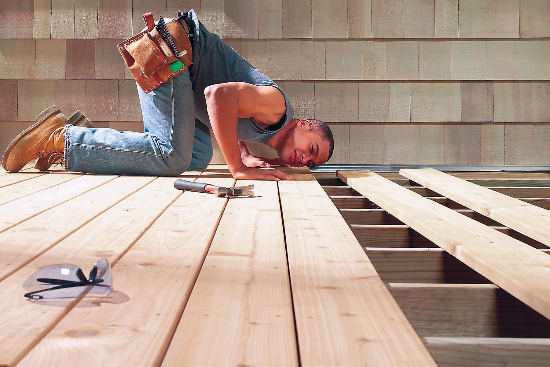 A person wearing jeans, a sleeveless shirt, and work boots kneels on a wooden deck under construction. They are looking closely at the wooden planks while holding a hammer. A tool belt is strapped to their waist, and safety glasses rest on the deck nearby.