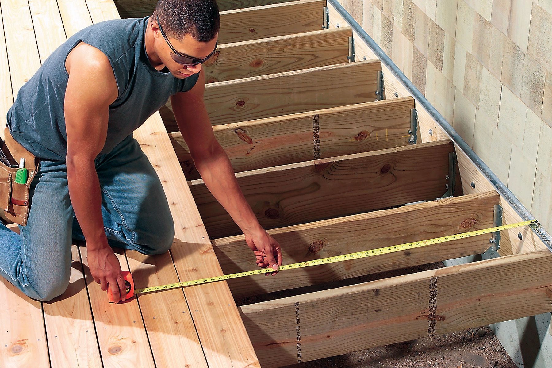 A person wearing safety glasses measures the width between wooden beams on a deck using a measuring tape. The deck is partially constructed, with several beams visible. The person is dressed in a sleeveless shirt and jeans, kneeling on one of the beams.