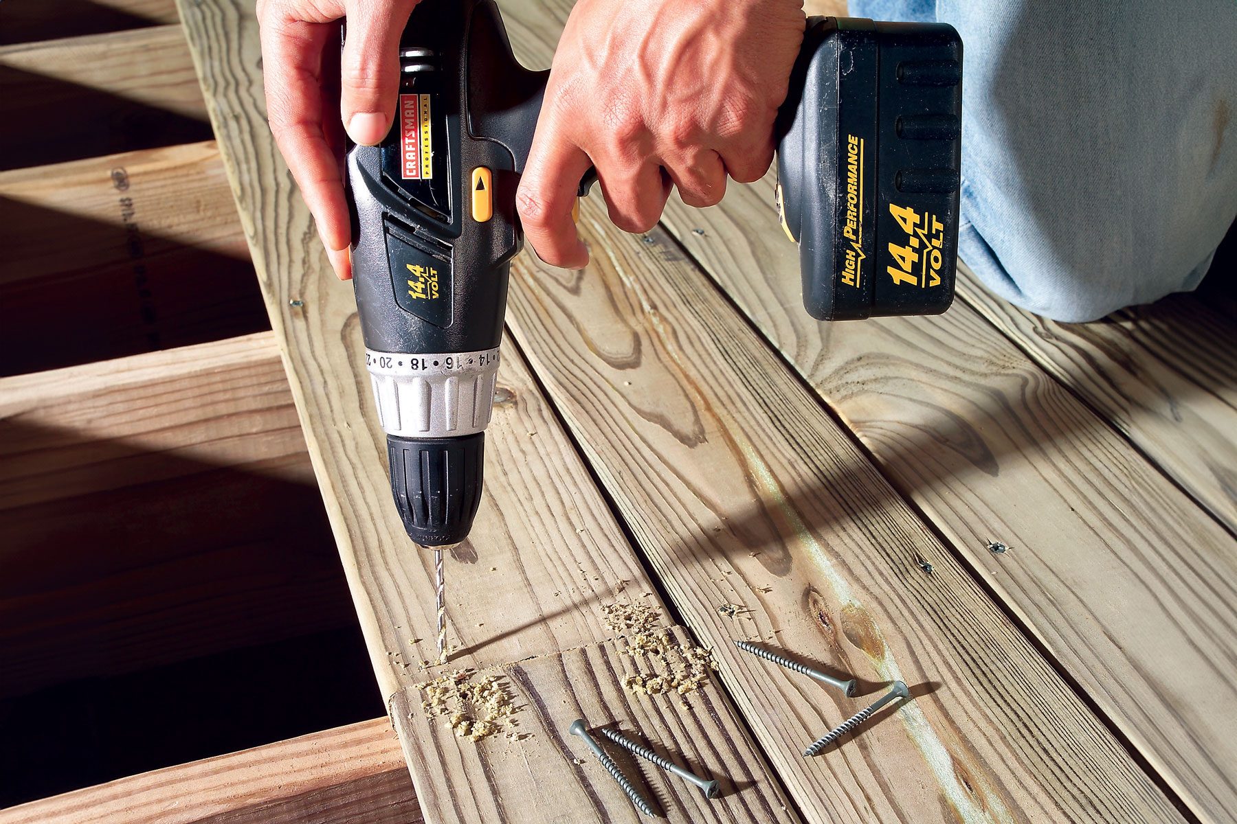 A person using a power drill to create a hole in a wooden plank. Several screws are lying on the plank beside the drilled holes..