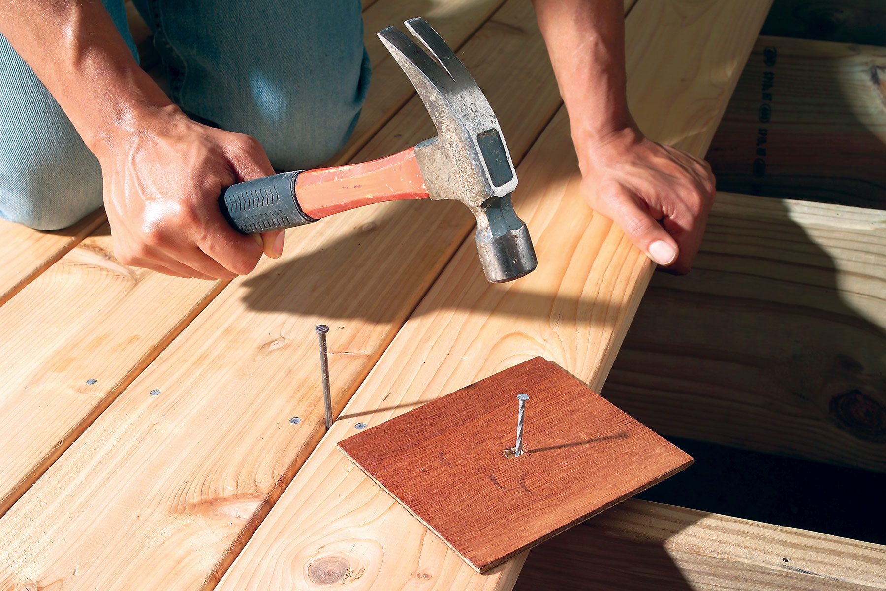 A person hammers a large nail into a wooden plank on a deck. They use a small square piece of wood to protect the plank's surface. The person’s hands and the hammer are visible, along with other wooden planks in the background.