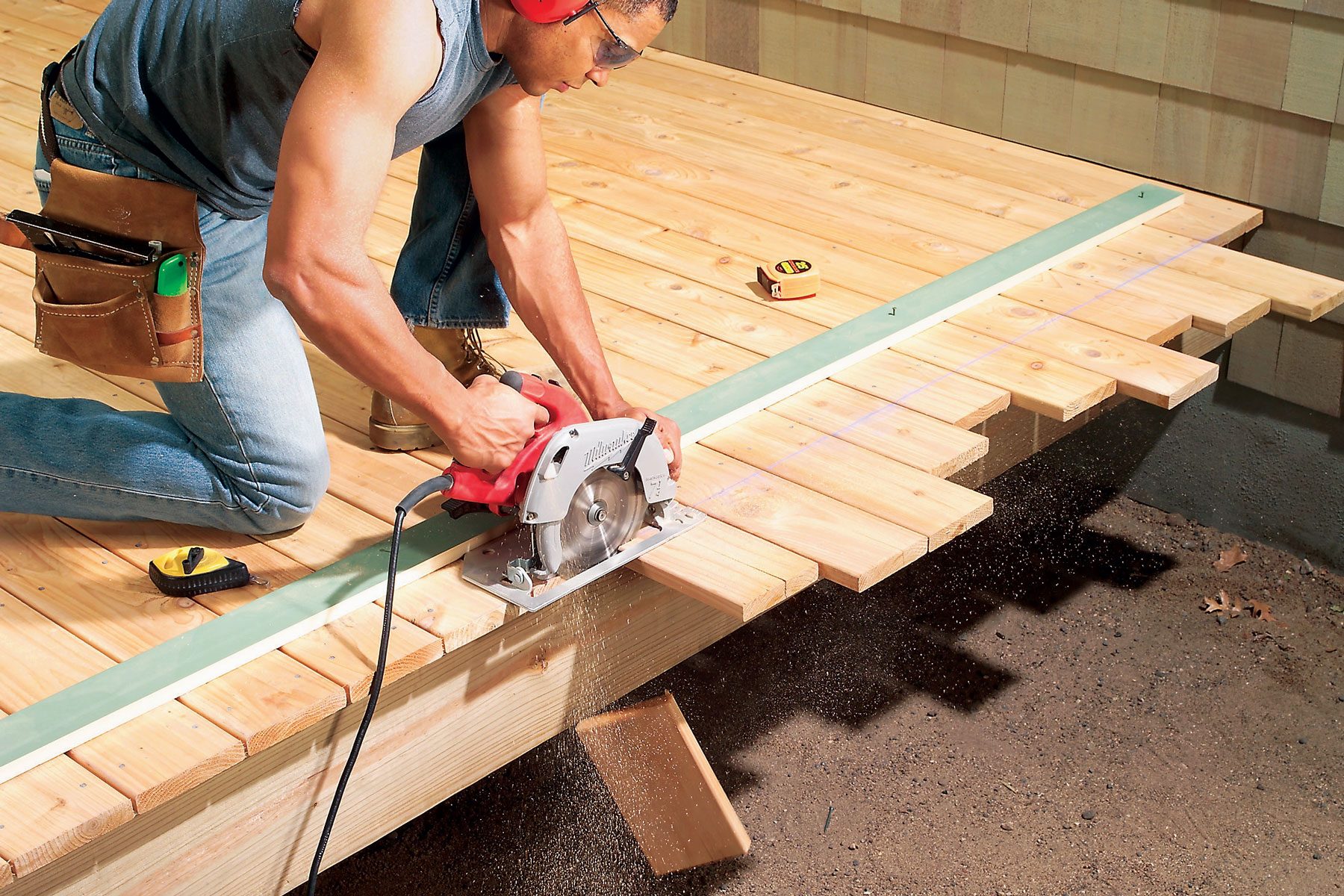 A person uses a circular saw to cut the edges of wooden planks on a deck. A measuring tape and pencil lines are visible, and sawdust is flying from the cutting area.