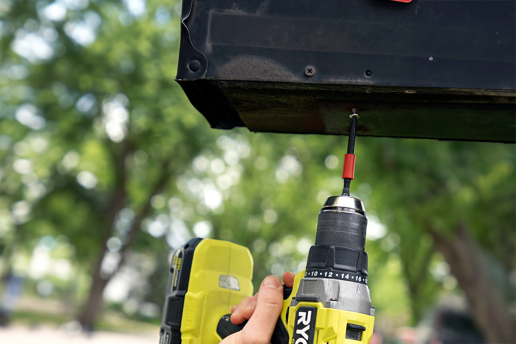 A close-up of a person using a yellow Ryobi power drill to drive a screw into a black metal surface. The background is blurred, showing some green trees, indicating an outdoor setting.