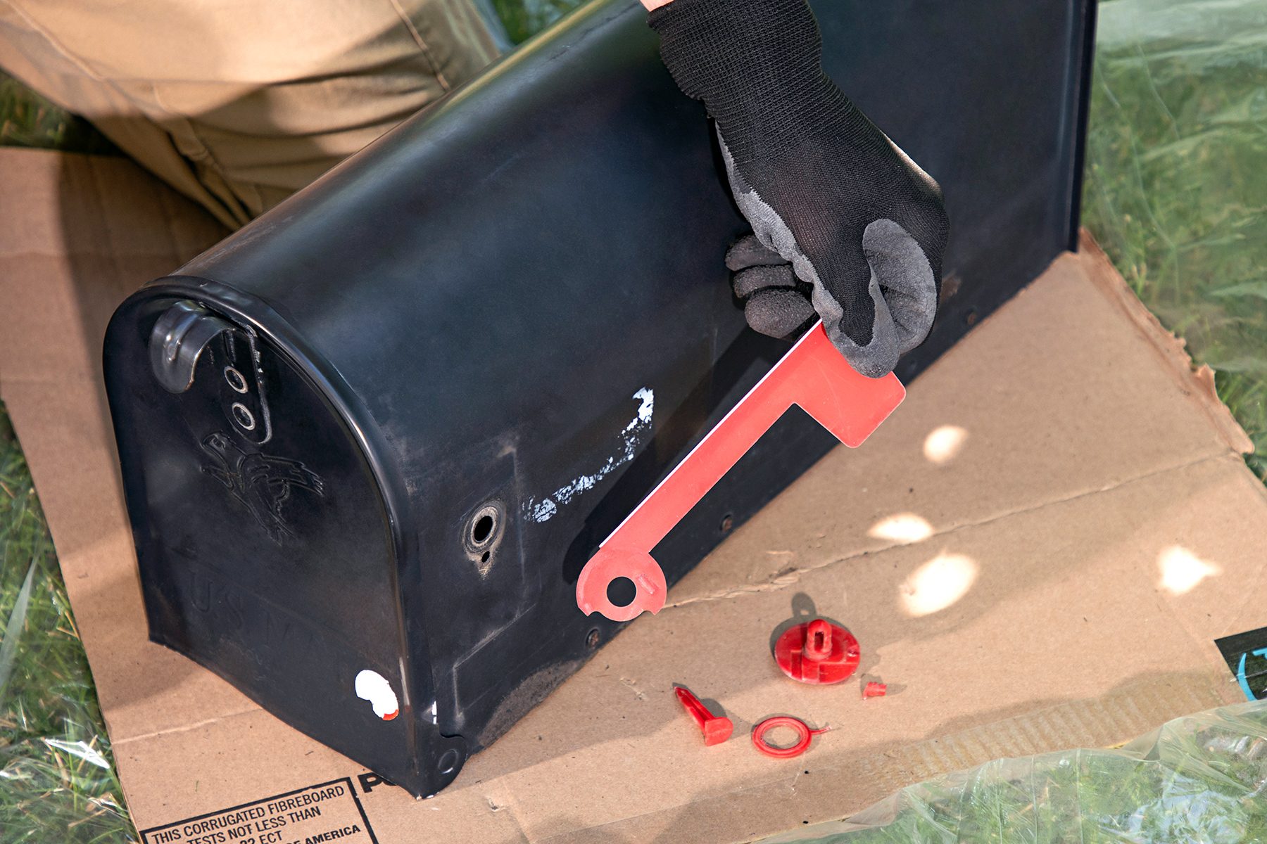 A person wearing a black glove is assembling a black mailbox on a cardboard surface. They are using a red tool, and there are small red components scattered nearby. The person is wearing beige pants, and grass is visible in the background.