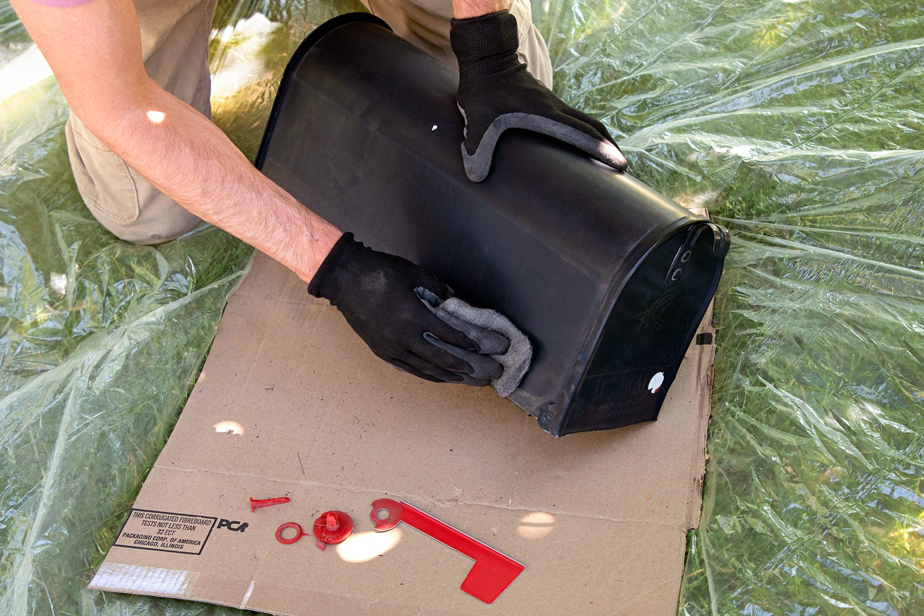 A person wearing black gloves is sanding a black mailbox placed on a piece of cardboard. The person is kneeling on what appears to be a plastic sheet covering grass. Red plastic components rest on the cardboard near the mailbox.