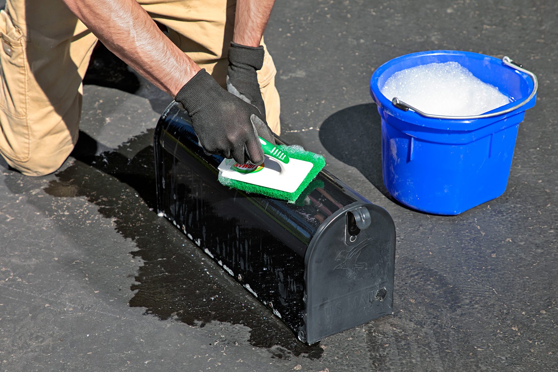 A person wearing gloves is kneeling on the ground and scrubbing a black mailbox with a sponge. Next to them is a blue bucket filled with soapy water.