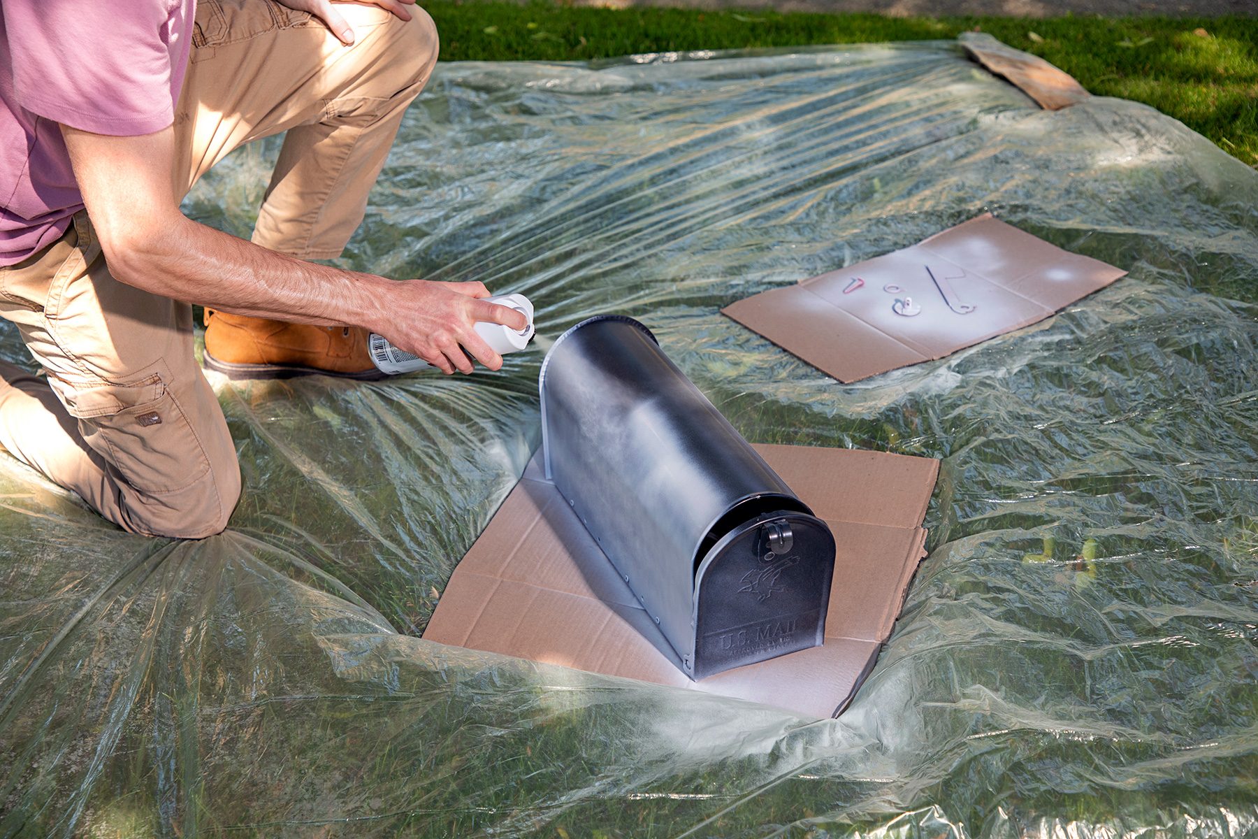 A person is spray painting a black mailbox placed on a piece of cardboard. The mailbox is set on a plastic sheet spread over the grass. The person is kneeling and wearing beige pants and a pink shirt.